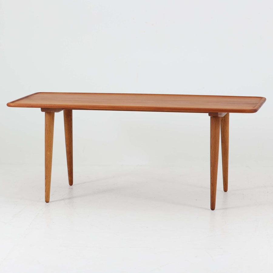 Hans J. Wegner（ハンス・J・ウェグナー）によるAndreas Tuck社製のセンターテーブル「AT11」です。幅130cmの広々とした天板はチーク無垢材を使用し、縁には繊細な面取り加工が施され、柔らかな印象を与えます。脚部には堅牢なオーク無垢材が用いられ、軽やかなテーパードデザインが北欧家具らしいバランスの良さを感じさせます。さらに脚のネジ部分まで木製で仕上げられており、細部に至るまで職人技が光る逸品です。脚は取り外し可能な構造で、輸送や設置も容易です。シンプルでありながら素材の美しさが際立ち、経年変化による深みを増した木肌がビンテージならではの存在感を放ちます。リビングの中心に置いて使うことで、空間に上質な雰囲気を添える一台です。