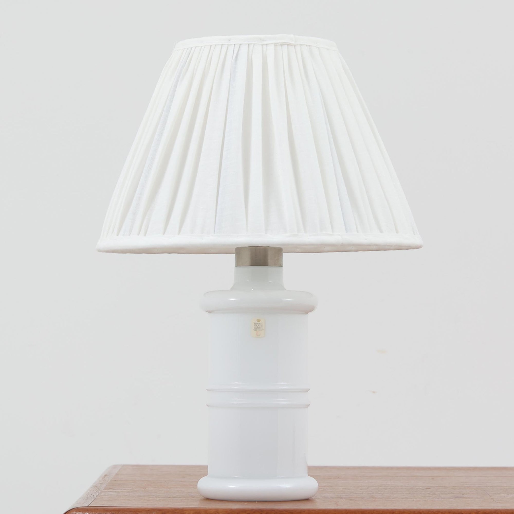 Sidse Wernerがデザインし、Holmegaard社より製造された「Apoteker」テーブルランプは、高さ46cmの堂々としたサイズ感と、白色ガラスの透明感が魅力の一品です。シリンダー状のフォルムにさりげなく入ったライン装飾が、シンプルながらも奥行きを感じさせるデザインとなっています。ガラス特有の柔らかな光の反射とリネンシェードを透過する穏やかな灯りは、リビングや寝室に落ち着いた雰囲気をもたらします。北欧ビンテージならではの洗練された存在感を楽しめる逸品です。