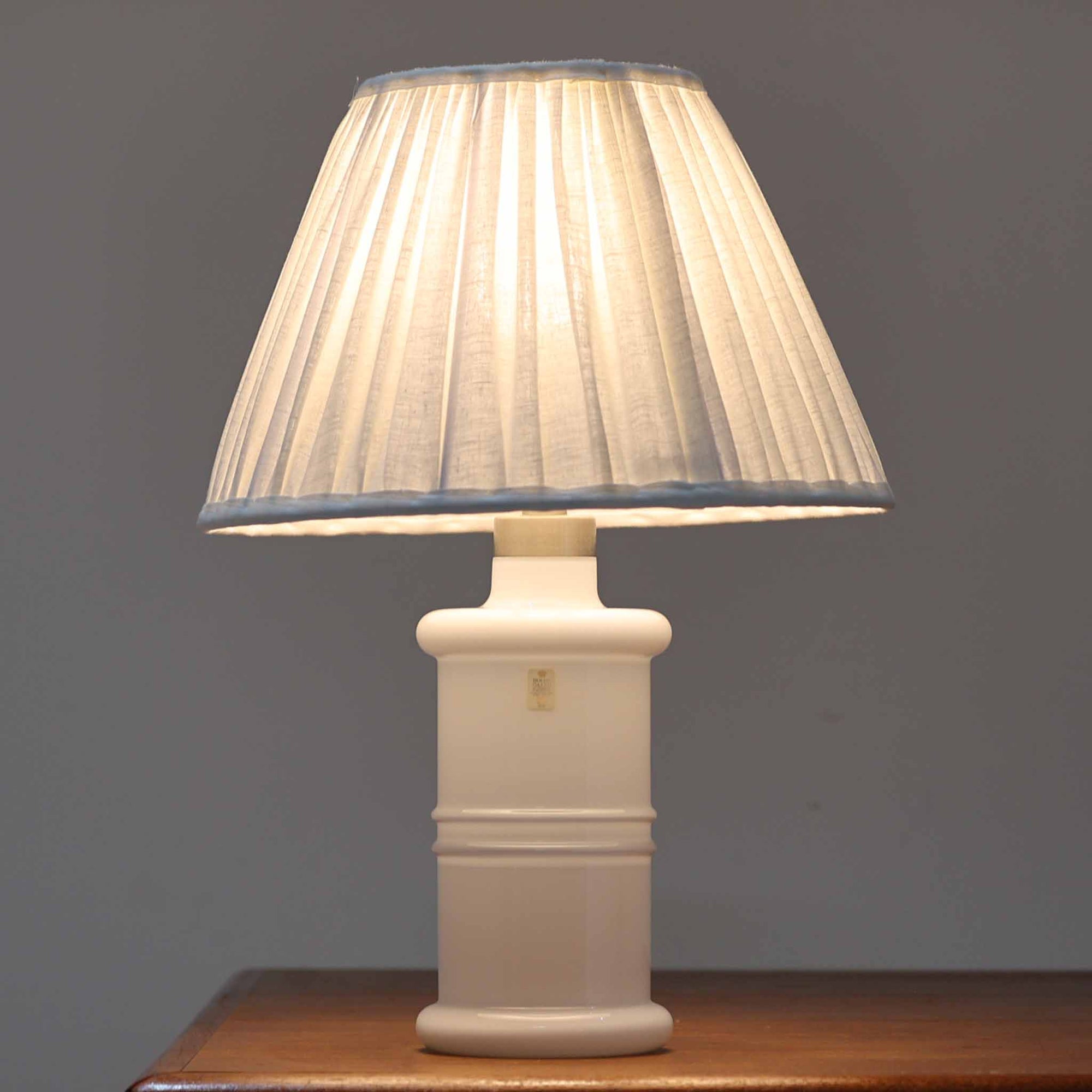 Sidse Wernerがデザインし、Holmegaard社より製造された「Apoteker」テーブルランプは、高さ46cmの堂々としたサイズ感と、白色ガラスの透明感が魅力の一品です。シリンダー状のフォルムにさりげなく入ったライン装飾が、シンプルながらも奥行きを感じさせるデザインとなっています。ガラス特有の柔らかな光の反射とリネンシェードを透過する穏やかな灯りは、リビングや寝室に落ち着いた雰囲気をもたらします。北欧ビンテージならではの洗練された存在感を楽しめる逸品です。