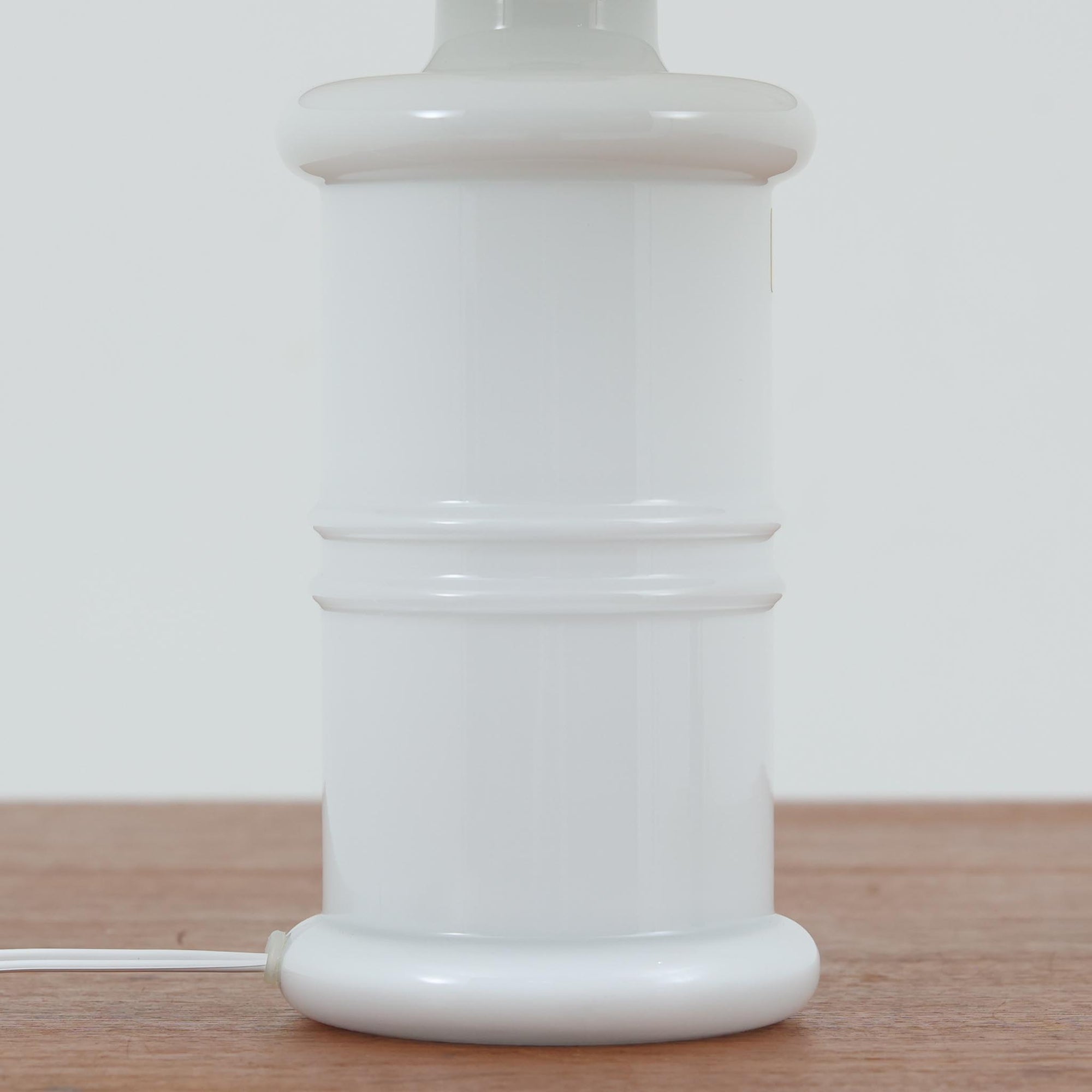 Sidse Wernerがデザインし、Holmegaard社より製造された「Apoteker」テーブルランプは、高さ46cmの堂々としたサイズ感と、白色ガラスの透明感が魅力の一品です。シリンダー状のフォルムにさりげなく入ったライン装飾が、シンプルながらも奥行きを感じさせるデザインとなっています。ガラス特有の柔らかな光の反射とリネンシェードを透過する穏やかな灯りは、リビングや寝室に落ち着いた雰囲気をもたらします。北欧ビンテージならではの洗練された存在感を楽しめる逸品です。