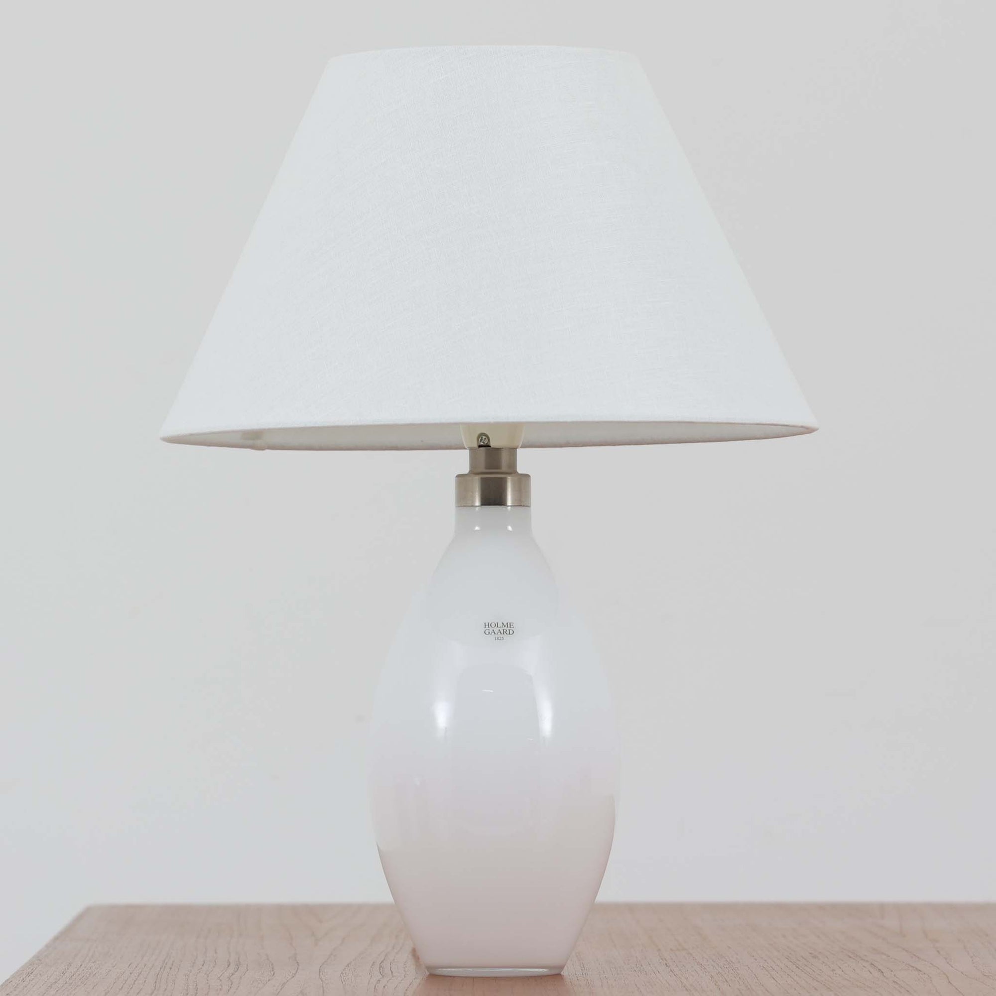 デンマークの老舗ガラスメーカー、Holmegaard（ホルムガード）社による「Cocoon」モデルのテーブルランプです。デザイナーはPeter Svarrer（ピーター・スヴァレ）で、彼らしい滑らかな曲線と透明感のあるフォルムが美しく表現されています。乳白色のガラスベースは光をやわらかく反射し、リネンシェードを通して穏やかな灯りを広げます。北欧らしいミニマルで洗練されたデザインは、ベッドサイドやリビングなどに上質な雰囲気をもたらしてくれます。