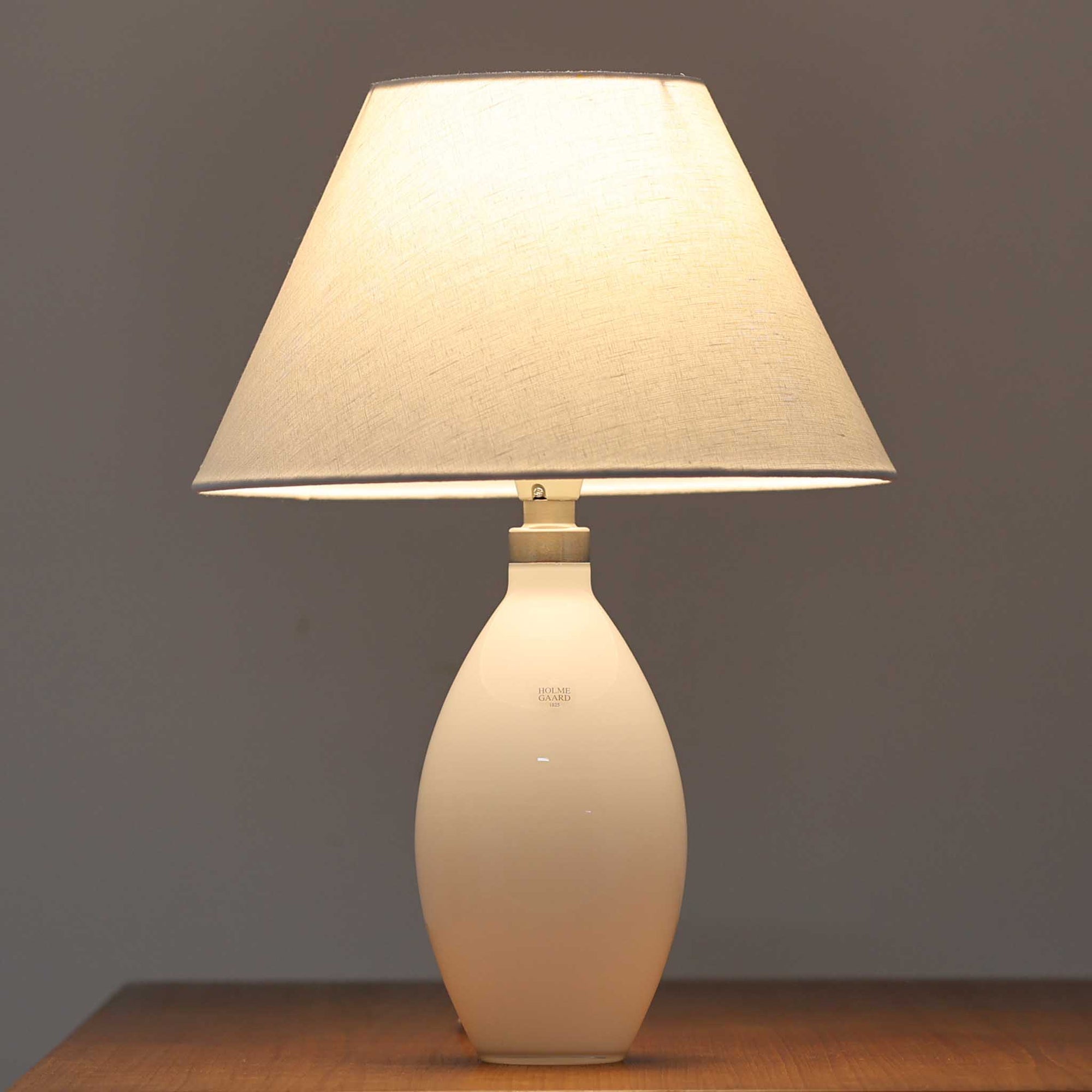 デンマークの老舗ガラスメーカー、Holmegaard（ホルムガード）社による「Cocoon」モデルのテーブルランプです。デザイナーはPeter Svarrer（ピーター・スヴァレ）で、彼らしい滑らかな曲線と透明感のあるフォルムが美しく表現されています。乳白色のガラスベースは光をやわらかく反射し、リネンシェードを通して穏やかな灯りを広げます。北欧らしいミニマルで洗練されたデザインは、ベッドサイドやリビングなどに上質な雰囲気をもたらしてくれます。