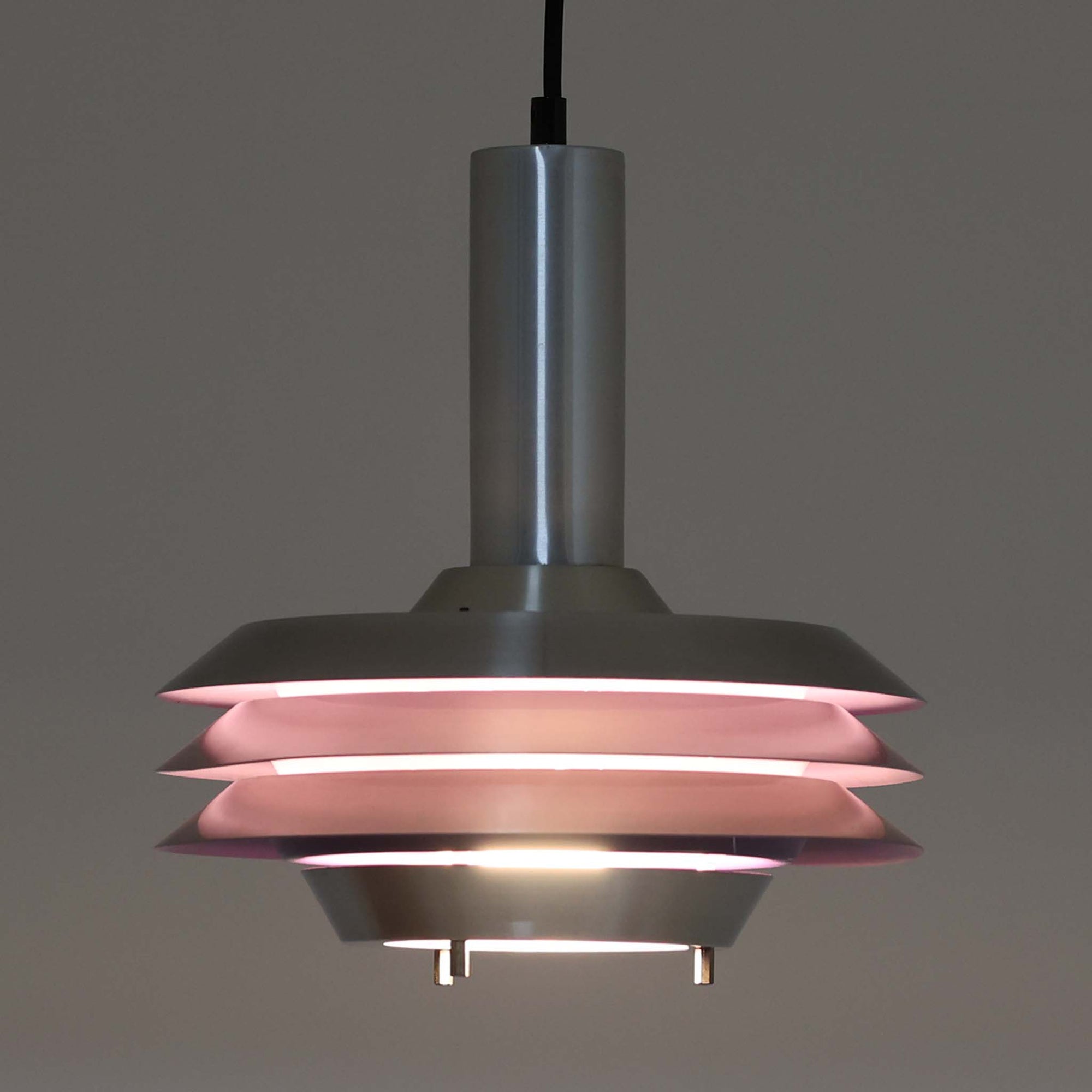 Carl Thoreがデザインし、Granhaga社より発表された直径28cmのペンダントライトです。アルミ製のシェードを幾重にも重ねた構造が特徴で、点灯すると隙間から柔らかな光が漏れ、独特の陰影と奥行きを生み出します。外側はシルバー、内側には紫がかった色味が施され、消灯時と点灯時で異なる表情を楽しめます。モダンでありながら温かみのある北欧らしい光のデザインは、ダイニングやリビングの照明として空間にアクセントを与えます。ビンテージならではの存在感を放つ希少な一品です。