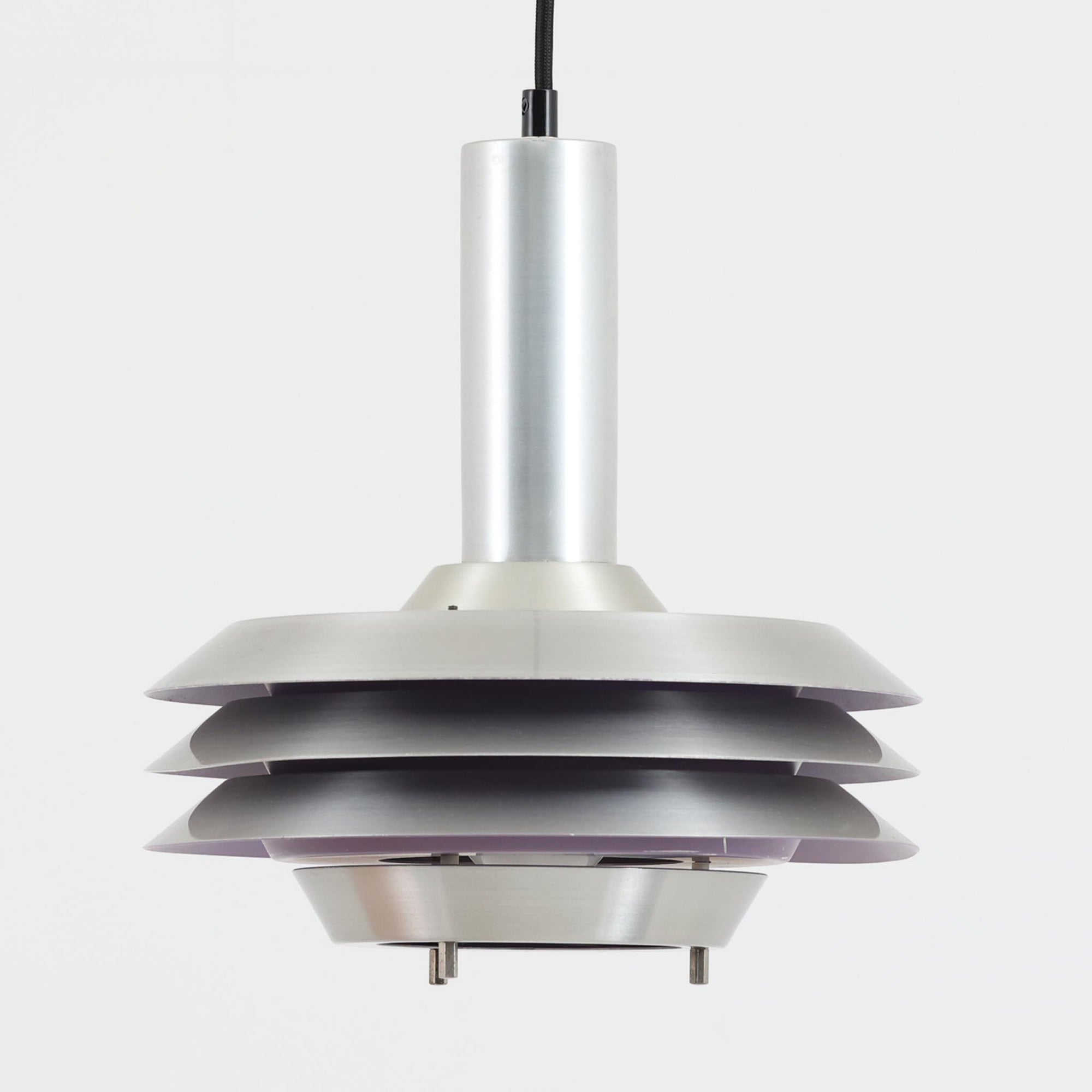 Carl Thoreがデザインし、Granhaga社より発表された直径28cmのペンダントライトです。アルミ製のシェードを幾重にも重ねた構造が特徴で、点灯すると隙間から柔らかな光が漏れ、独特の陰影と奥行きを生み出します。外側はシルバー、内側には紫がかった色味が施され、消灯時と点灯時で異なる表情を楽しめます。モダンでありながら温かみのある北欧らしい光のデザインは、ダイニングやリビングの照明として空間にアクセントを与えます。ビンテージならではの存在感を放つ希少な一品です。