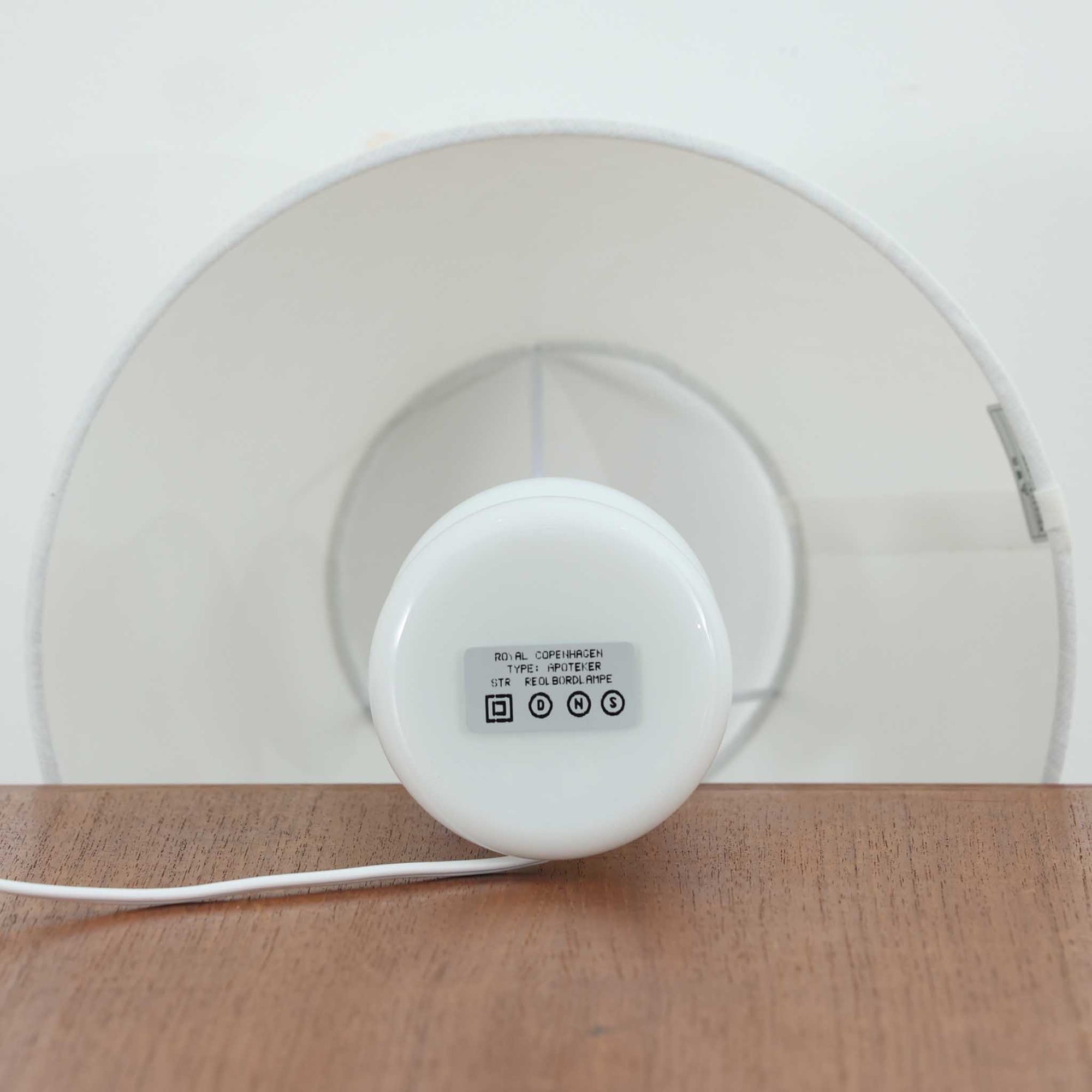 Sidse Werner（シセ・ヴェルナー）がデザインし、Holmegaard（ホルムガード）より製造されたテーブルランプ「Apoteker」です。高さ29cmと小ぶりなサイズで、艶やかなホワイトガラスのベースが柔らかな印象を与えます。リネンシェードを通した光は優しく拡散し、ナチュラルな雰囲気を演出。シンプルでありながら彫刻的なフォルムが存在感を放ち、ベッドサイドやデスク、リビングのサイドテーブルなど様々な空間で活躍します。北欧デザインの温もりを感じられるビンテージ照明です。