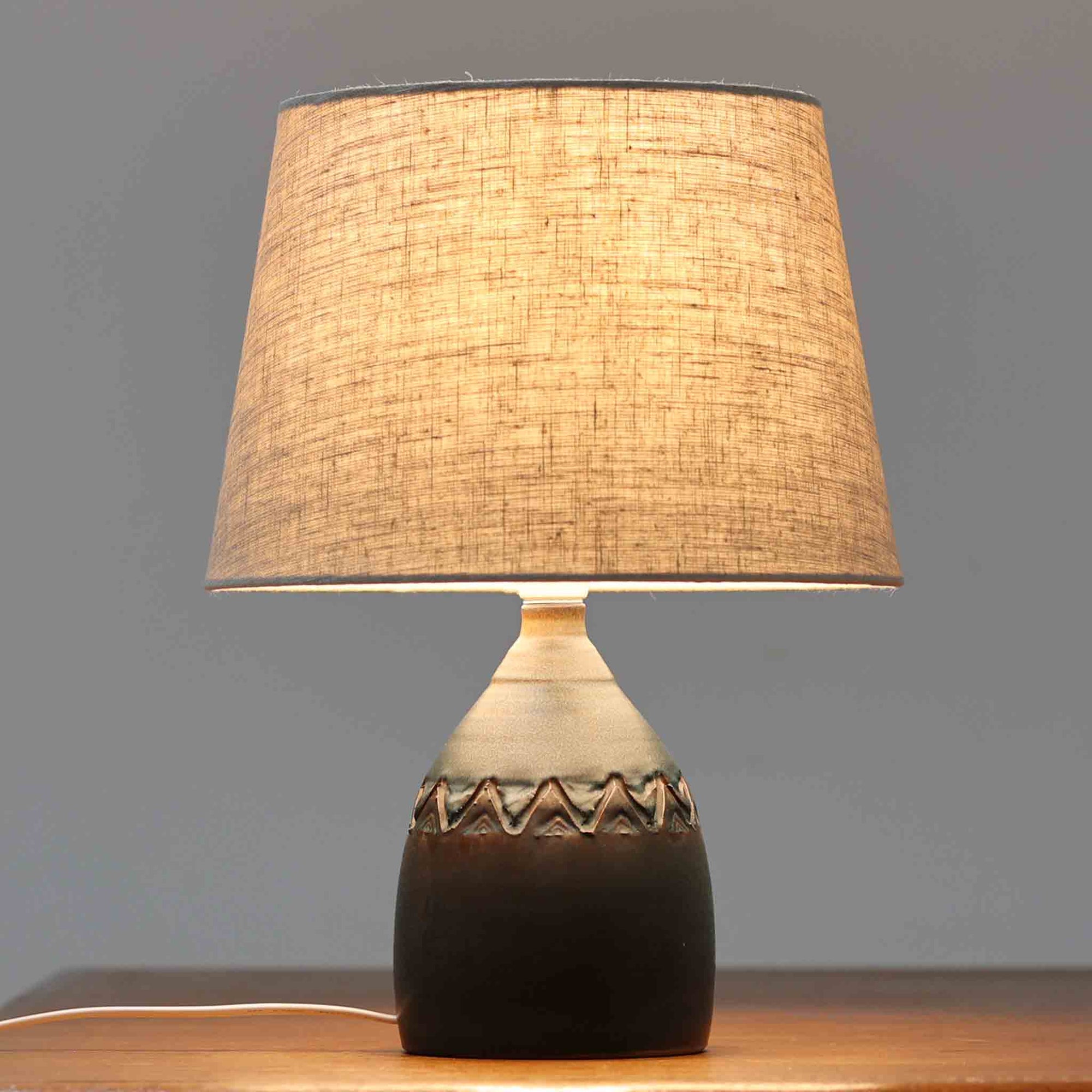デンマーク製のテーブルランプは、高さ36cmとコンパクトで扱いやすく、陶磁器ベースには深みのある釉薬と素朴な質感が調和しています。胴部には幾何学的なレリーフが施され、落ち着いた佇まいの中に個性が光ります。リネンシェードを透過する柔らかな光が空間に温もりを与え、リビングや寝室のアクセントとしても最適です。メーカー不詳ながら、スタジオポタリーならではの職人技と一点物の味わいを感じられる北欧ビンテージ品です。