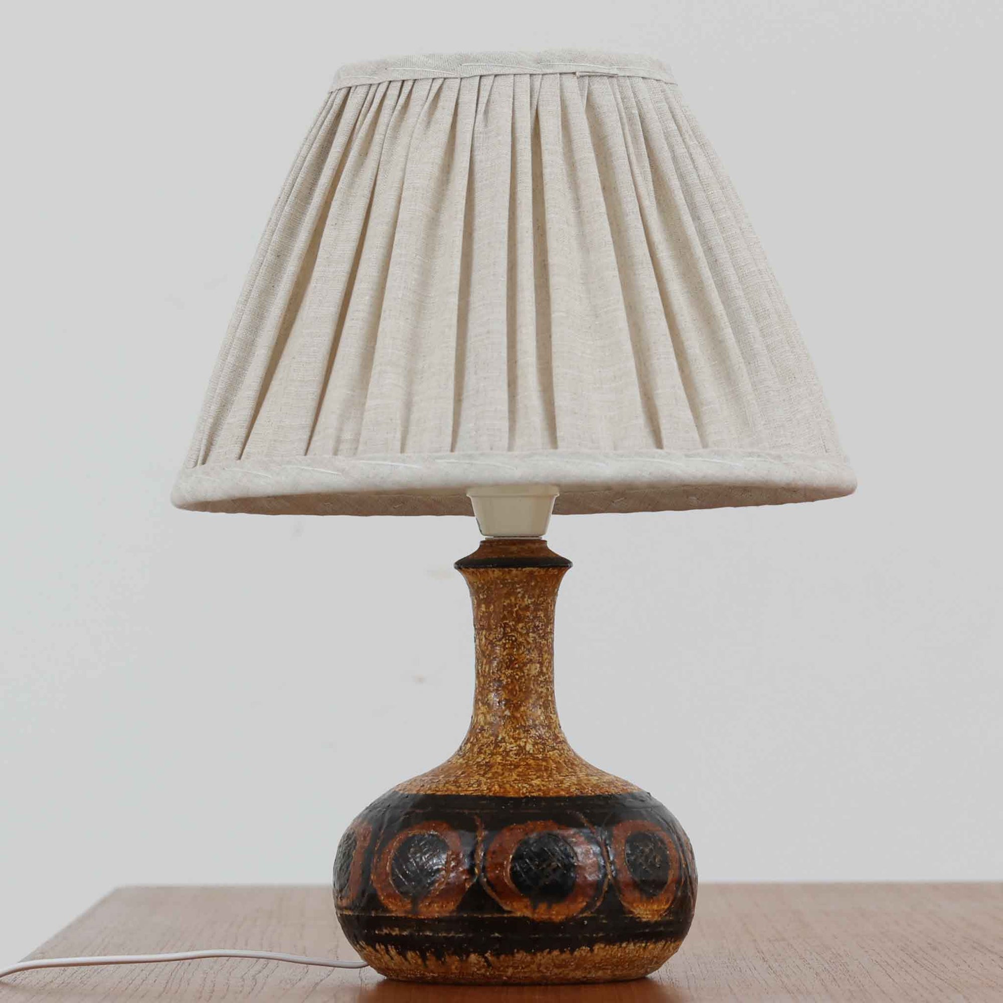 デンマークの陶芸家 Jette Helleroe（イェッテ・ヘレロー）によるテーブルランプです。素朴な質感と温かみのある釉薬のグラデーションが魅力で、ベースには深みのあるブラウンとオレンジを基調とした幾何学的な模様が施されています。手仕事ならではの味わいが感じられ、点灯時にはやわらかな灯りが陶器の表情をより一層引き立てます。ナチュラルなリネンシェードとの組み合わせが優しく、北欧らしい落ち着いた雰囲気を空間にもたらします。