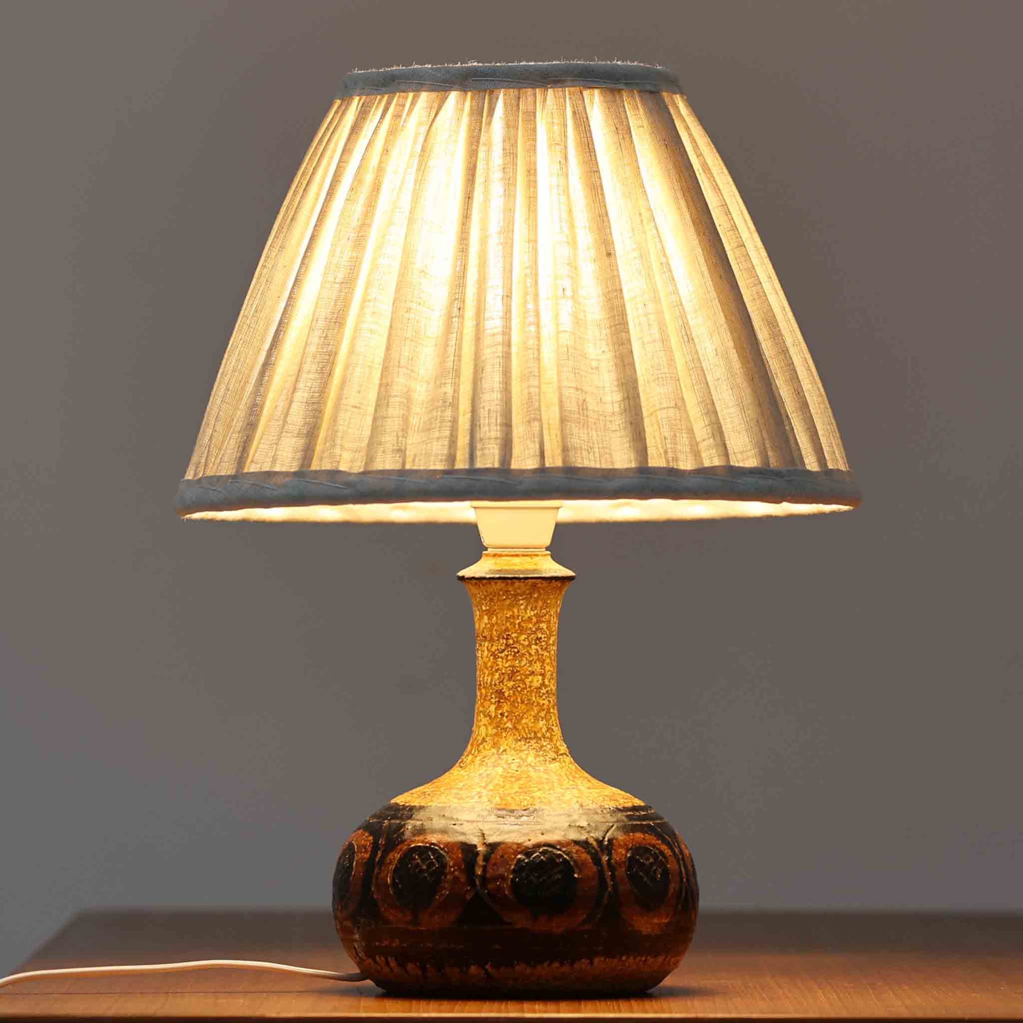 デンマークの陶芸家 Jette Helleroe（イェッテ・ヘレロー）によるテーブルランプです。素朴な質感と温かみのある釉薬のグラデーションが魅力で、ベースには深みのあるブラウンとオレンジを基調とした幾何学的な模様が施されています。手仕事ならではの味わいが感じられ、点灯時にはやわらかな灯りが陶器の表情をより一層引き立てます。ナチュラルなリネンシェードとの組み合わせが優しく、北欧らしい落ち着いた雰囲気を空間にもたらします。