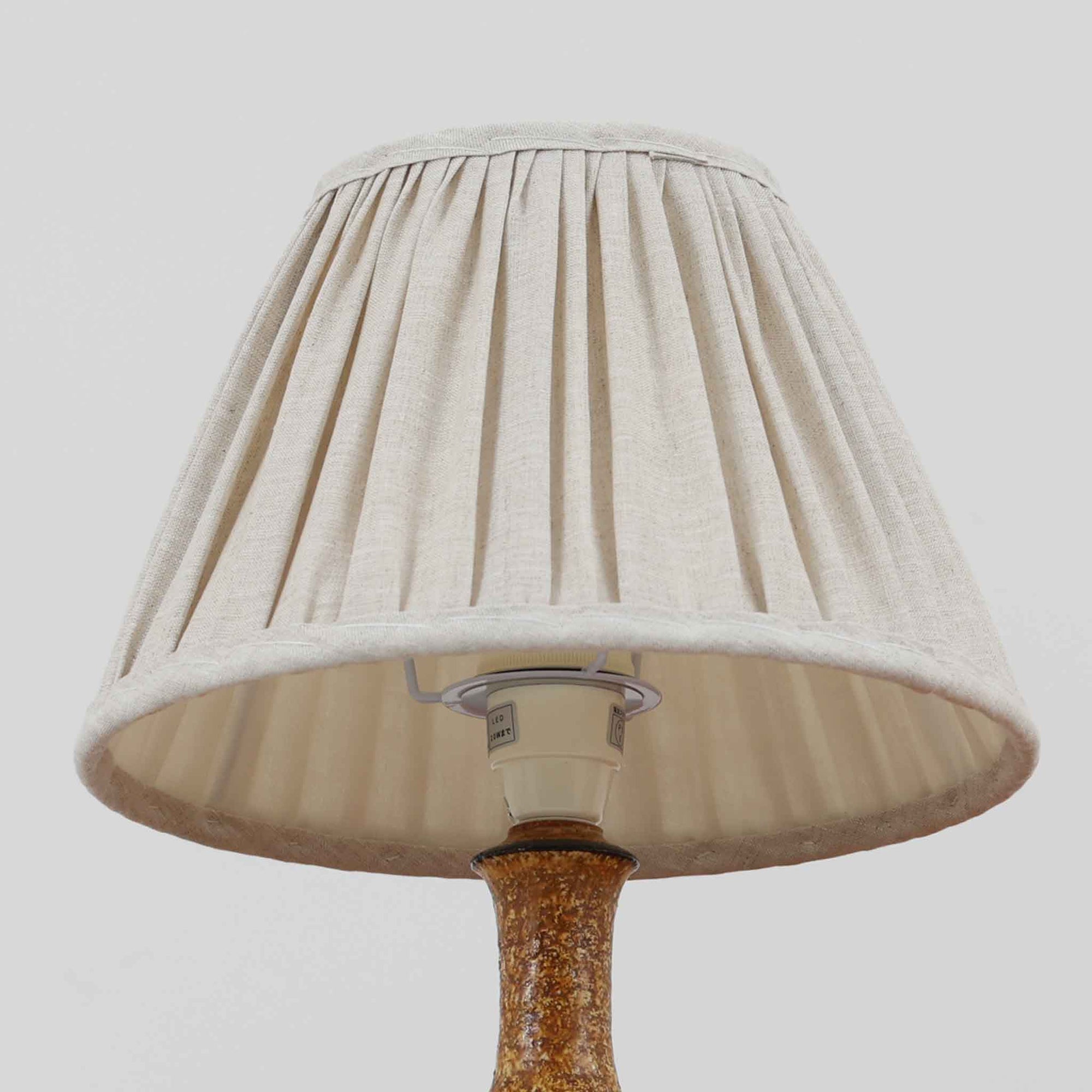 デンマークの陶芸家 Jette Helleroe（イェッテ・ヘレロー）によるテーブルランプです。素朴な質感と温かみのある釉薬のグラデーションが魅力で、ベースには深みのあるブラウンとオレンジを基調とした幾何学的な模様が施されています。手仕事ならではの味わいが感じられ、点灯時にはやわらかな灯りが陶器の表情をより一層引き立てます。ナチュラルなリネンシェードとの組み合わせが優しく、北欧らしい落ち着いた雰囲気を空間にもたらします。