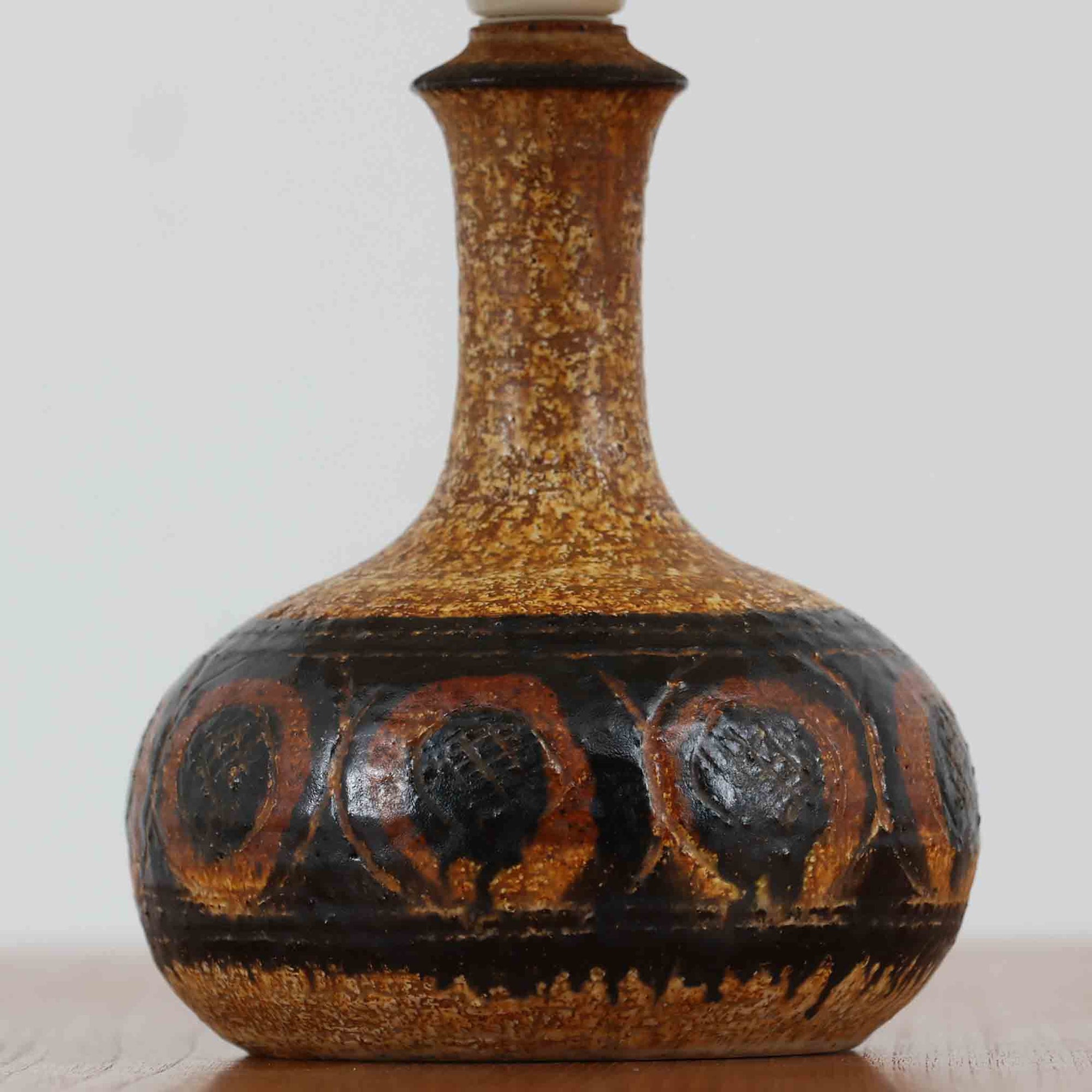 デンマークの陶芸家 Jette Helleroe（イェッテ・ヘレロー）によるテーブルランプです。素朴な質感と温かみのある釉薬のグラデーションが魅力で、ベースには深みのあるブラウンとオレンジを基調とした幾何学的な模様が施されています。手仕事ならではの味わいが感じられ、点灯時にはやわらかな灯りが陶器の表情をより一層引き立てます。ナチュラルなリネンシェードとの組み合わせが優しく、北欧らしい落ち着いた雰囲気を空間にもたらします。