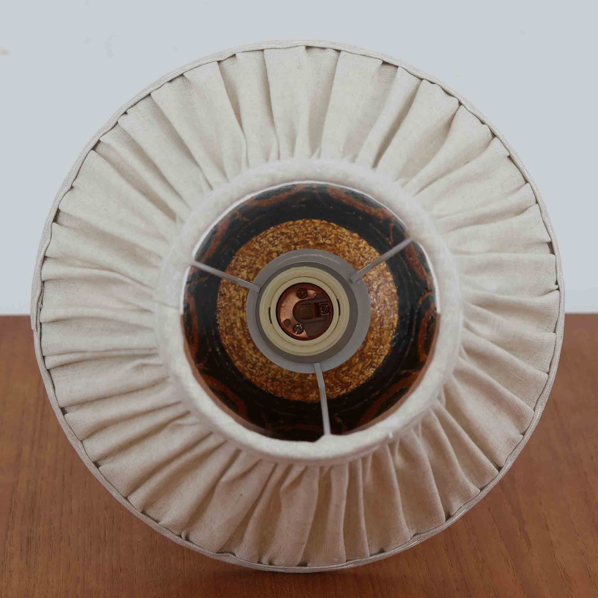 デンマークの陶芸家 Jette Helleroe（イェッテ・ヘレロー）によるテーブルランプです。素朴な質感と温かみのある釉薬のグラデーションが魅力で、ベースには深みのあるブラウンとオレンジを基調とした幾何学的な模様が施されています。手仕事ならではの味わいが感じられ、点灯時にはやわらかな灯りが陶器の表情をより一層引き立てます。ナチュラルなリネンシェードとの組み合わせが優しく、北欧らしい落ち着いた雰囲気を空間にもたらします。