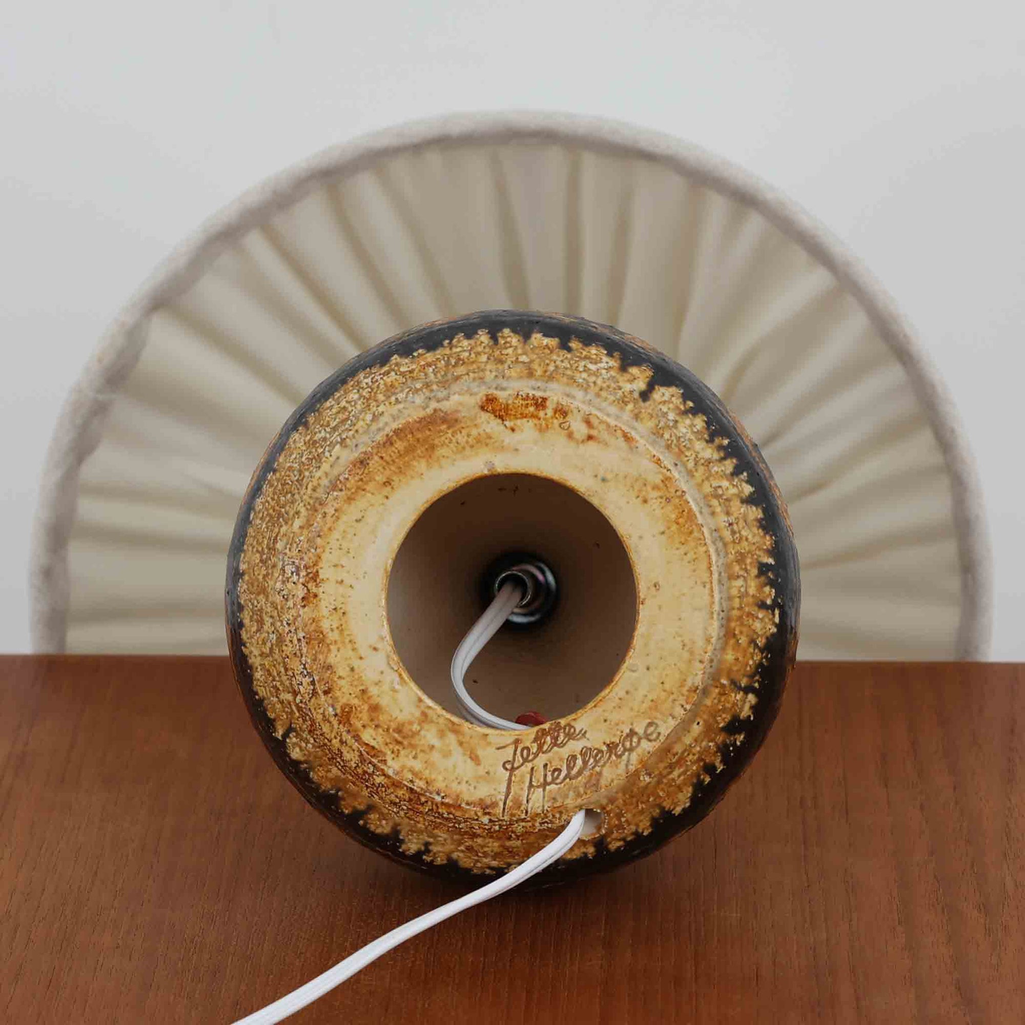 デンマークの陶芸家 Jette Helleroe（イェッテ・ヘレロー）によるテーブルランプです。素朴な質感と温かみのある釉薬のグラデーションが魅力で、ベースには深みのあるブラウンとオレンジを基調とした幾何学的な模様が施されています。手仕事ならではの味わいが感じられ、点灯時にはやわらかな灯りが陶器の表情をより一層引き立てます。ナチュラルなリネンシェードとの組み合わせが優しく、北欧らしい落ち着いた雰囲気を空間にもたらします。