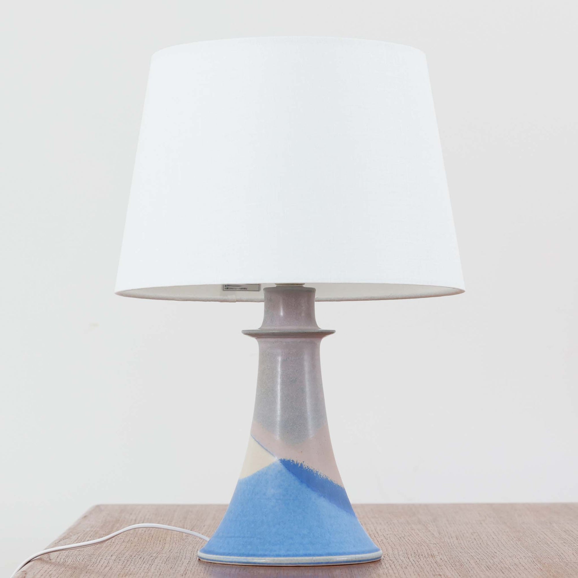 デンマーク製、高さ40cmのテーブルランプです。陶磁器製のベースはブルーとベージュが美しく溶け合うグラデーションが特徴で、ハンドクラフトならではの味わい深い質感が魅力です。裏面には「J.H」の刻印があり、デザインや色使いからデンマークの陶芸家 Jette Hellerøe（イェッテ・ヘレロー）の作品である可能性が高いものの、確証はございません。シンプルなリネンシェードを通した柔らかな光が落ち着いた雰囲気を演出し、リビングやベッドサイドに温もりを添えます。北欧らしいナチュラルな美しさを感じさせるビンテージ照明で、インテリアのアクセントとしてもおすすめの一品です。