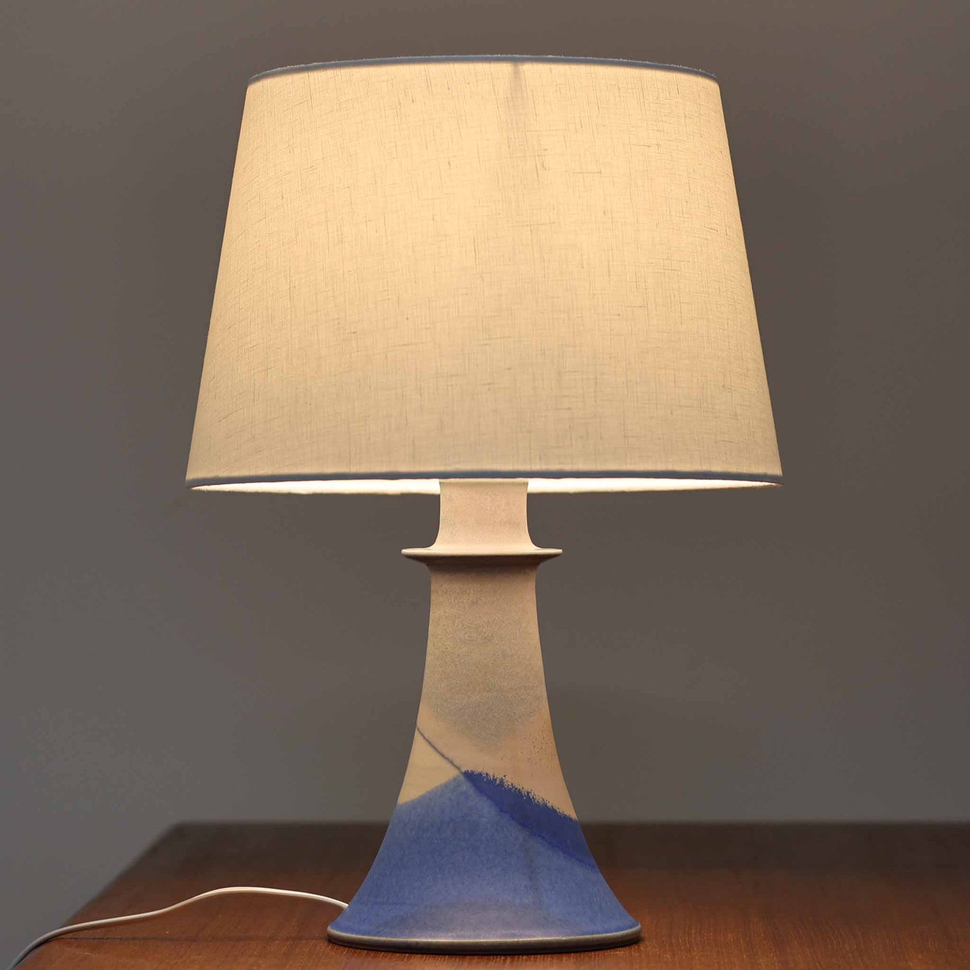 デンマーク製、高さ40cmのテーブルランプです。陶磁器製のベースはブルーとベージュが美しく溶け合うグラデーションが特徴で、ハンドクラフトならではの味わい深い質感が魅力です。裏面には「J.H」の刻印があり、デザインや色使いからデンマークの陶芸家 Jette Hellerøe（イェッテ・ヘレロー）の作品である可能性が高いものの、確証はございません。シンプルなリネンシェードを通した柔らかな光が落ち着いた雰囲気を演出し、リビングやベッドサイドに温もりを添えます。北欧らしいナチュラルな美しさを感じさせるビンテージ照明で、インテリアのアクセントとしてもおすすめの一品です。