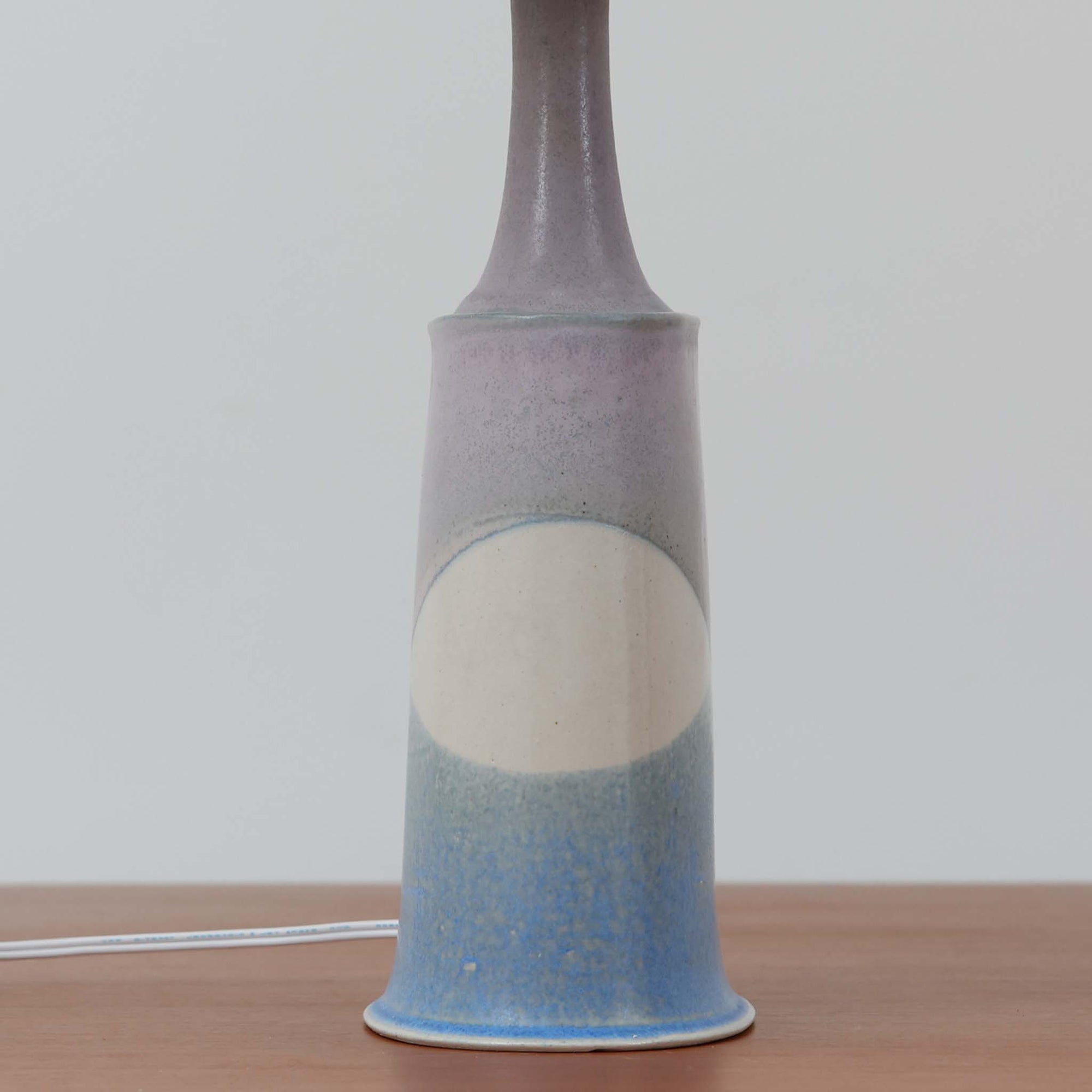 こちらのテーブルランプは、陶磁器ベースに淡いパープルやブルーを溶け合わせたような独特の釉薬表情が魅力で、ビンテージならではの柔らかなグラデーションが優しい存在感を放つ一品です。細く立ち上がる支柱にリネンシェードを組み合わせたシンプルな構造は安定感があり、点灯時にはシェード越しに温かく穏やかな光が広がります。高さ46cmと扱いやすいサイズで、ベッドサイドやリビングのサイドテーブルにも取り入れやすく、北欧らしい静かな色彩と経年の味わいを楽しめるビンテージ照明です。