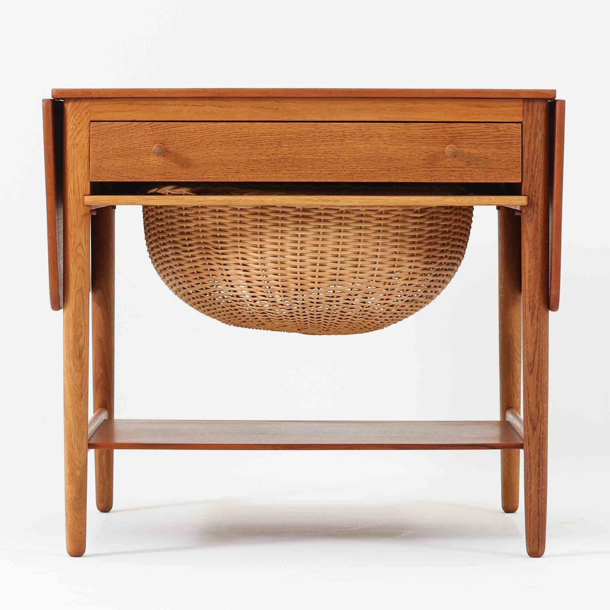 ハンス・J・ウェグナーが手がけ、Andreas Tuck社より製作されたAT33ソーイングテーブルは、天板を広げられる独創的な構造と実用性を兼ね備えた北欧ビンテージを代表する一台です。中央には引き出しと仕切り付き収納、下部にはラタンバスケットを備え、道具をすっきりと収められる設計が魅力。AT33は複数の素材構成で展開されたモデルですが、こちらは天板にチーク突板材、脚部にオーク無垢材を用いた仕様で、素材のコントラストと軽やかな佇まいが特徴です。異素材を巧みに組み合わせた構造と端正なプロポーションは、使うほどに職人技と経年変化の美しさを実感できます。