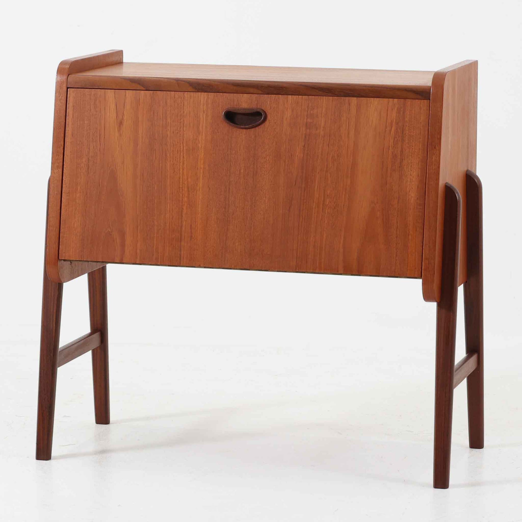 チーク材を用いた小ぶりなキャビネットは、直線を基調としつつ柔らかな丸みを持たせた側板と脚部のラインが印象的な北欧ビンテージ家具です。前板一枚で構成されたフラップ扉には掘り込み式の取っ手が設けられ、開閉時の所作も美しく、内部には実用的な収納棚を備えています。幅69cmと取り入れやすいサイズ感で、リビングや玄関、ベッドサイドの収納としても活躍し、経年により深みを増したチーク材の木目と職人技が静かな存在感を放つ一台です。