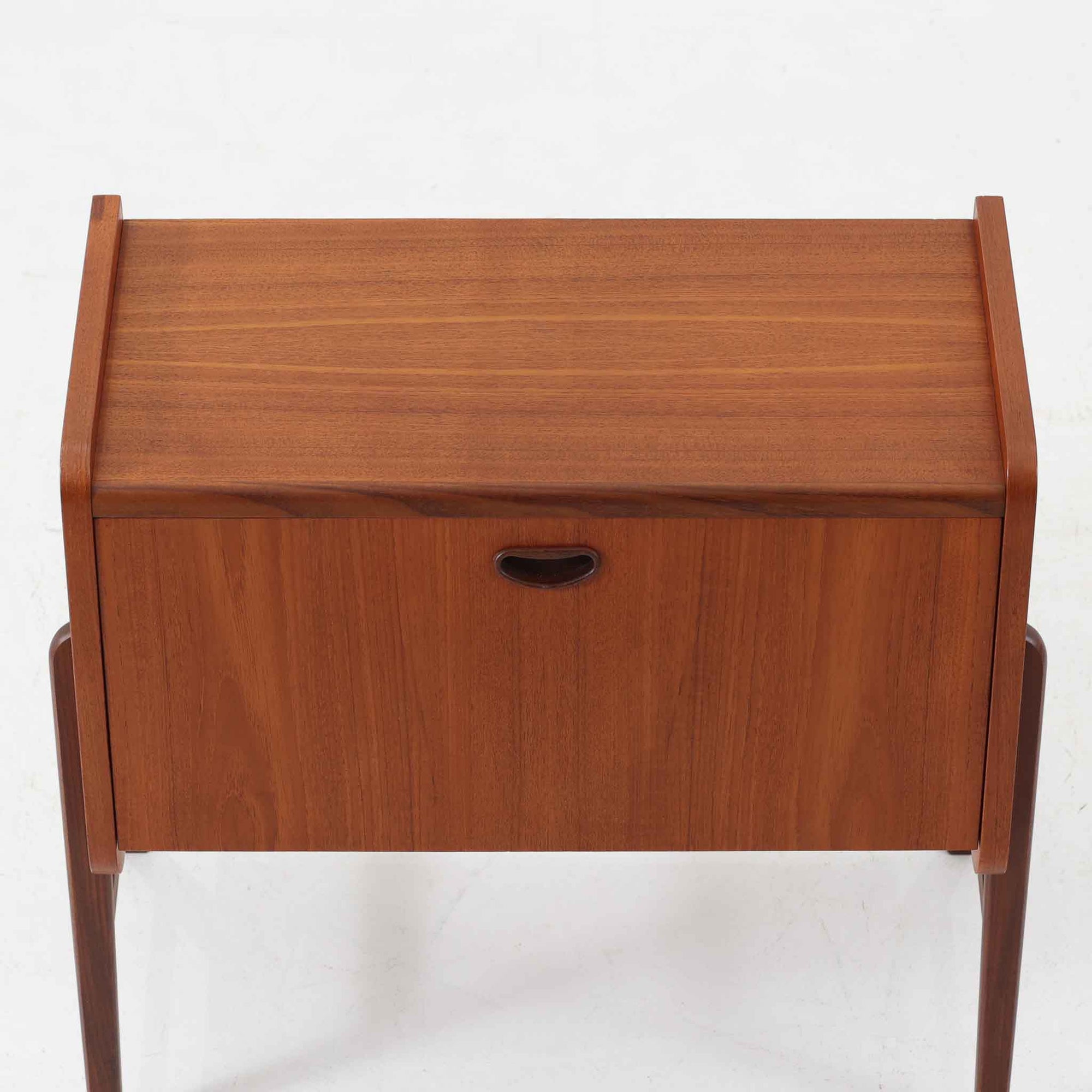 チーク材を用いた小ぶりなキャビネットは、直線を基調としつつ柔らかな丸みを持たせた側板と脚部のラインが印象的な北欧ビンテージ家具です。前板一枚で構成されたフラップ扉には掘り込み式の取っ手が設けられ、開閉時の所作も美しく、内部には実用的な収納棚を備えています。幅69cmと取り入れやすいサイズ感で、リビングや玄関、ベッドサイドの収納としても活躍し、経年により深みを増したチーク材の木目と職人技が静かな存在感を放つ一台です。