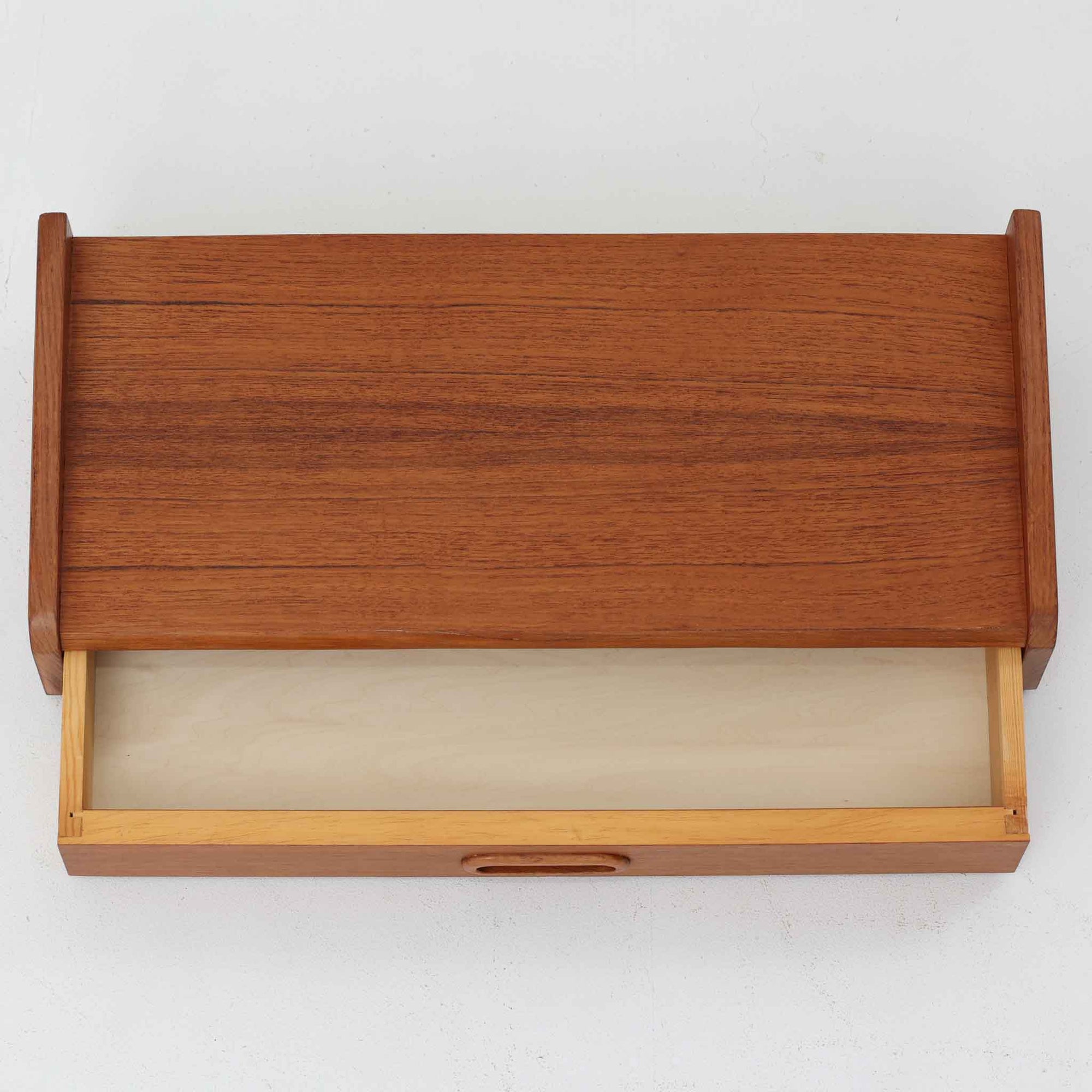 チーク材を用いたこちらのウォールシェルフは、直線的な棚板に両端の立ち上がりを設けた端正なフォルムが印象的な北欧ビンテージ家具です。前面には掘り込み式の取っ手を備えた引き出しが組み込まれており、見た目の軽やかさを保ちながら、実用的な収納力も確保しています。壁面を有効活用できる構造のため、デスク上やベッドサイド、玄関周りの小物収納としても取り入れやすく、側板は縁に向かって面取りが施され、厚みの変化によって立体感と柔らかな印象を与えています。経年により深みを増したチーク材の木目と丁寧な作りが、空間に静かな存在感を添える一台です。