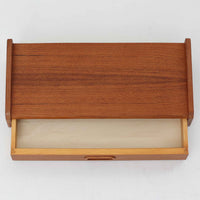 チーク材を用いたこちらのウォールシェルフは、直線的な棚板に両端の立ち上がりを設けた端正なフォルムが印象的な北欧ビンテージ家具です。前面には掘り込み式の取っ手を備えた引き出しが組み込まれており、見た目の軽やかさを保ちながら、実用的な収納力も確保しています。壁面を有効活用できる構造のため、デスク上やベッドサイド、玄関周りの小物収納としても取り入れやすく、側板は縁に向かって面取りが施され、厚みの変化によって立体感と柔らかな印象を与えています。経年により深みを増したチーク材の木目と丁寧な作りが、空間に静かな存在感を添える一台です。