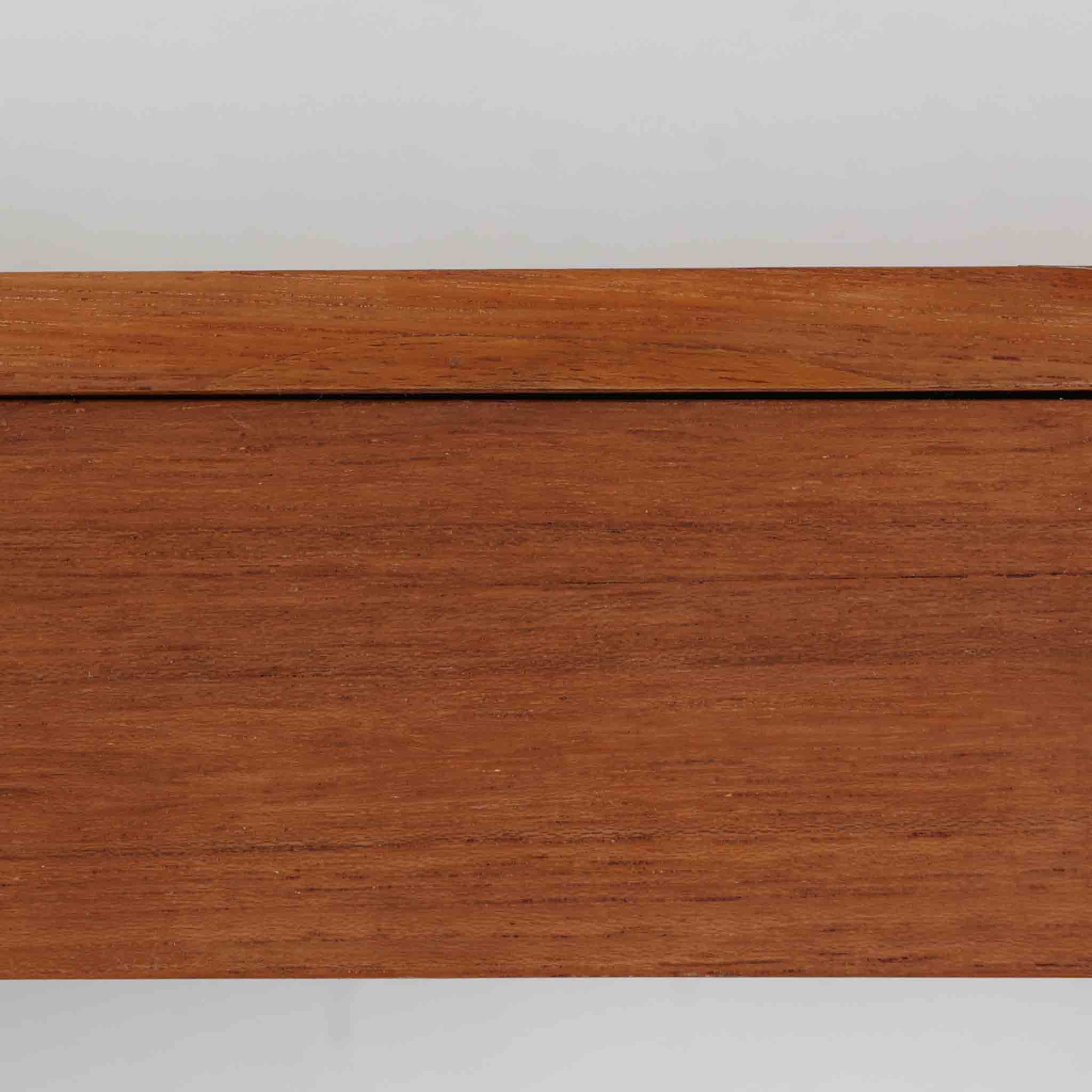 チーク材を用いたこちらのウォールシェルフは、直線的な棚板に両端の立ち上がりを設けた端正なフォルムが印象的な北欧ビンテージ家具です。前面には掘り込み式の取っ手を備えた引き出しが組み込まれており、見た目の軽やかさを保ちながら、実用的な収納力も確保しています。壁面を有効活用できる構造のため、デスク上やベッドサイド、玄関周りの小物収納としても取り入れやすく、側板は縁に向かって面取りが施され、厚みの変化によって立体感と柔らかな印象を与えています。経年により深みを増したチーク材の木目と丁寧な作りが、空間に静かな存在感を添える一台です。