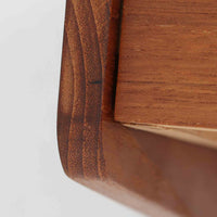 チーク材を用いたこちらのウォールシェルフは、直線的な棚板に両端の立ち上がりを設けた端正なフォルムが印象的な北欧ビンテージ家具です。前面には掘り込み式の取っ手を備えた引き出しが組み込まれており、見た目の軽やかさを保ちながら、実用的な収納力も確保しています。壁面を有効活用できる構造のため、デスク上やベッドサイド、玄関周りの小物収納としても取り入れやすく、側板は縁に向かって面取りが施され、厚みの変化によって立体感と柔らかな印象を与えています。経年により深みを増したチーク材の木目と丁寧な作りが、空間に静かな存在感を添える一台です。