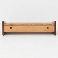 チーク材を用いたこちらのウォールシェルフは、直線的な棚板に両端の立ち上がりを設けた端正なフォルムが印象的な北欧ビンテージ家具です。前面には掘り込み式の取っ手を備えた引き出しが組み込まれており、見た目の軽やかさを保ちながら、実用的な収納力も確保しています。壁面を有効活用できる構造のため、デスク上やベッドサイド、玄関周りの小物収納としても取り入れやすく、側板は縁に向かって面取りが施され、厚みの変化によって立体感と柔らかな印象を与えています。経年により深みを増したチーク材の木目と丁寧な作りが、空間に静かな存在感を添える一台です。