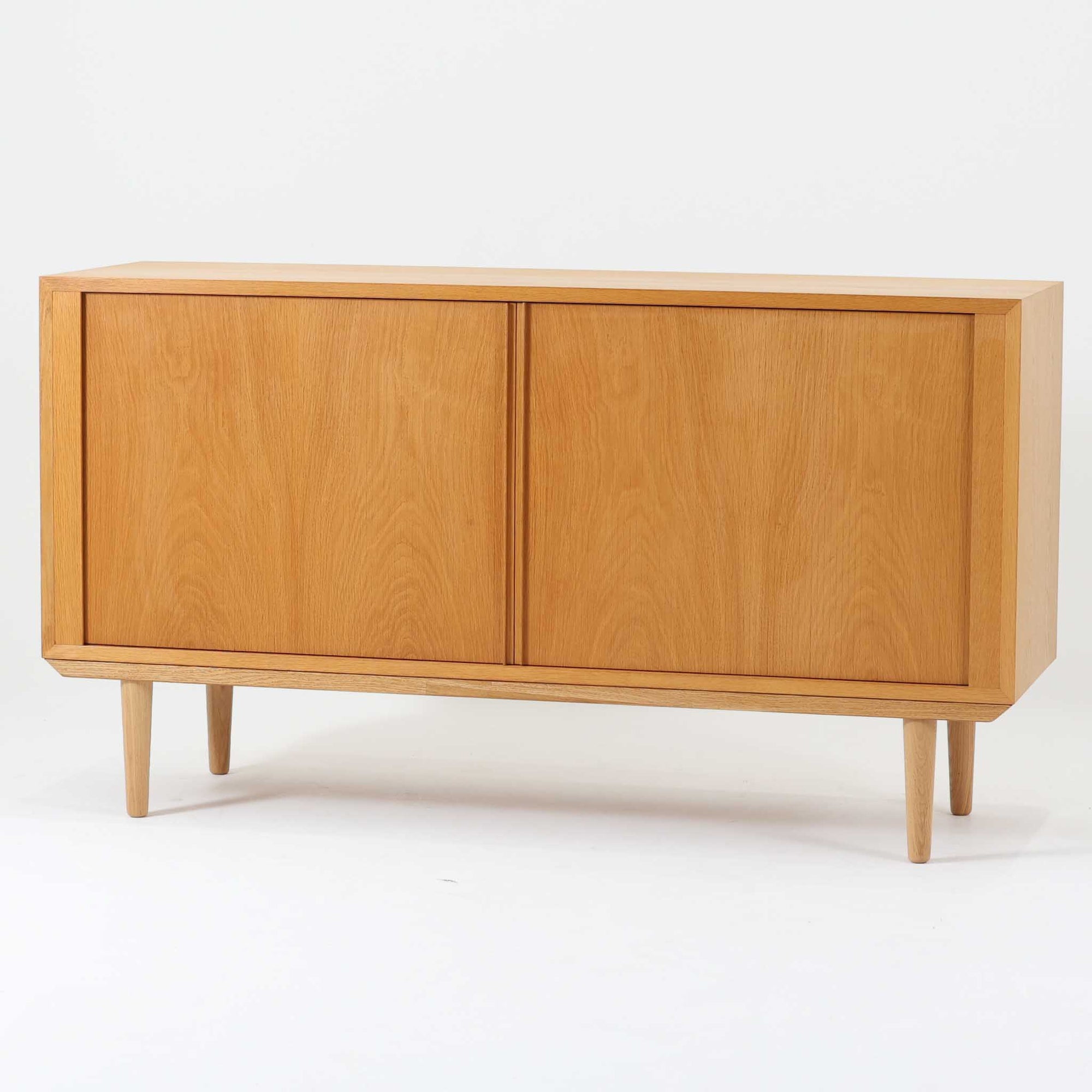 Poul Hundevadがデザインし、Hundevad & Coより製作された蛇腹仕様のサイドボードです。オーク材の穏やかで上品な木目を活かした外観に、横方向へ滑らかに開閉する蛇腹扉を組み合わせた、機能美あふれる一台。扉の開閉時に前後のスペースを取らない構造は、動線を妨げず実用性にも優れています。内部は棚板構成で収納力が高く、食器や書類、リビング周りの小物まで幅広く対応。細身の脚部が全体を軽やかに引き締め、経年によって深みを増した素材感が北欧ビンテージならではの静かな存在感を空間にもたらします。