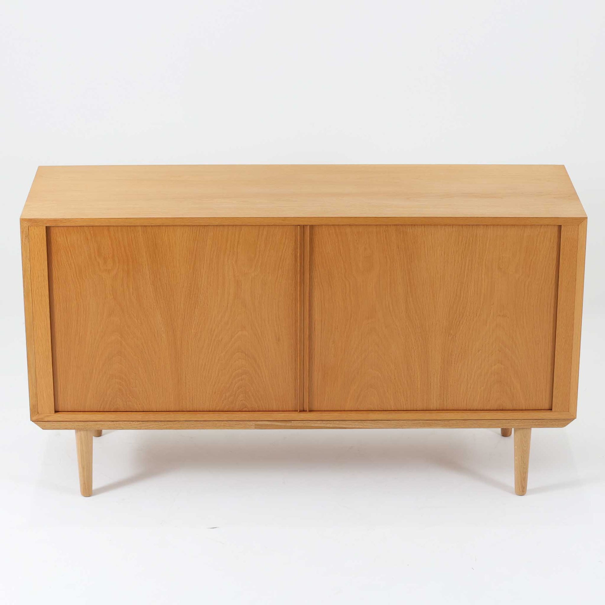 Poul Hundevadがデザインし、Hundevad &amp; Coより製作された蛇腹仕様のサイドボードです。オーク材の穏やかで上品な木目を活かした外観に、横方向へ滑らかに開閉する蛇腹扉を組み合わせた、機能美あふれる一台。扉の開閉時に前後のスペースを取らない構造は、動線を妨げず実用性にも優れています。内部は棚板構成で収納力が高く、食器や書類、リビング周りの小物まで幅広く対応。細身の脚部が全体を軽やかに引き締め、経年によって深みを増した素材感が北欧ビンテージならではの静かな存在感を空間にもたらします。