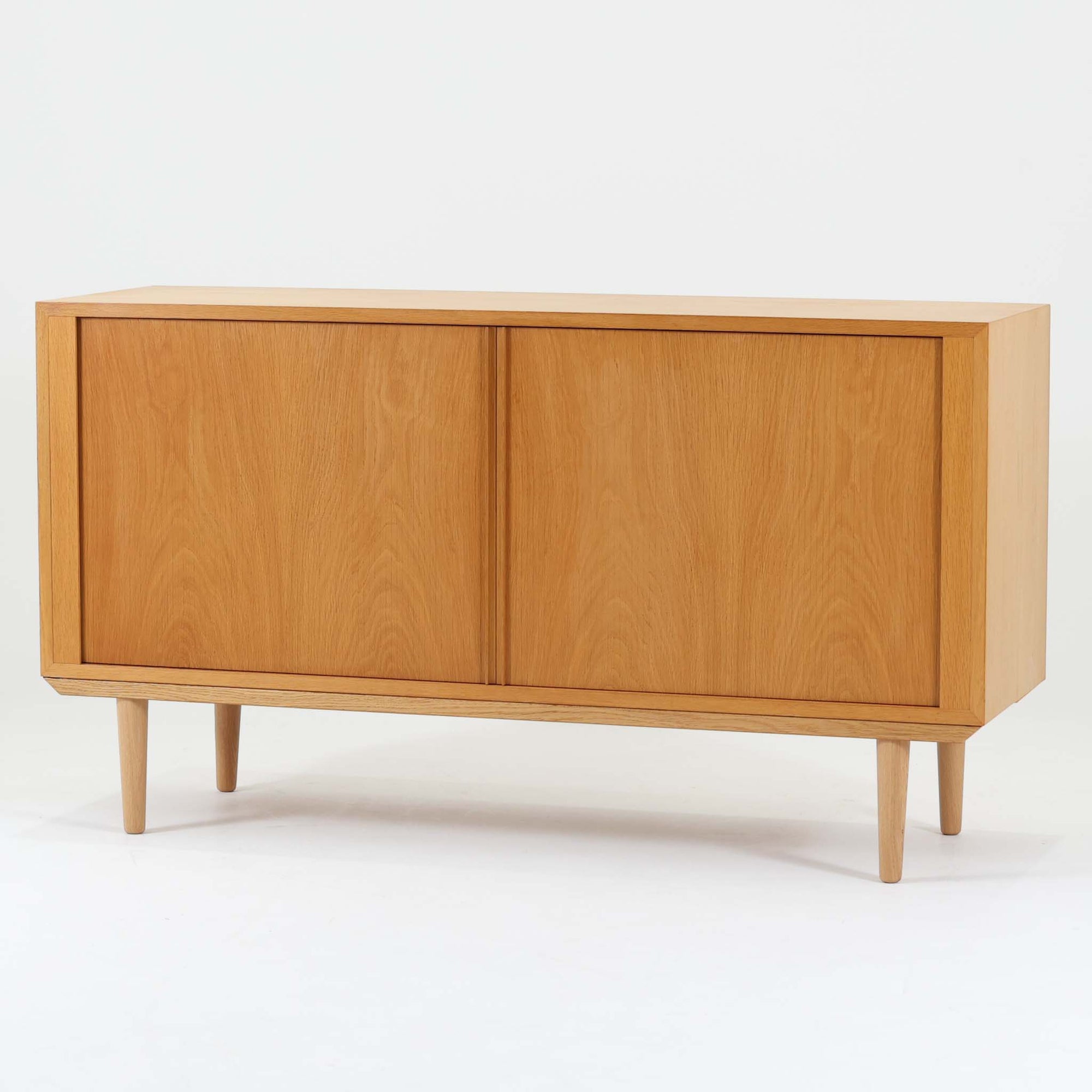 Poul Hundevadがデザインし、Hundevad & Coより製作された蛇腹仕様のサイドボードです。オーク材の穏やかで上品な木目を活かした外観に、横方向へ滑らかに開閉する蛇腹扉を組み合わせた、機能美あふれる一台。扉の開閉時に前後のスペースを取らない構造は、動線を妨げず実用性にも優れています。内部は棚板構成で収納力が高く、食器や書類、リビング周りの小物まで幅広く対応。細身の脚部が全体を軽やかに引き締め、経年によって深みを増した素材感が北欧ビンテージならではの静かな存在感を空間にもたらします。