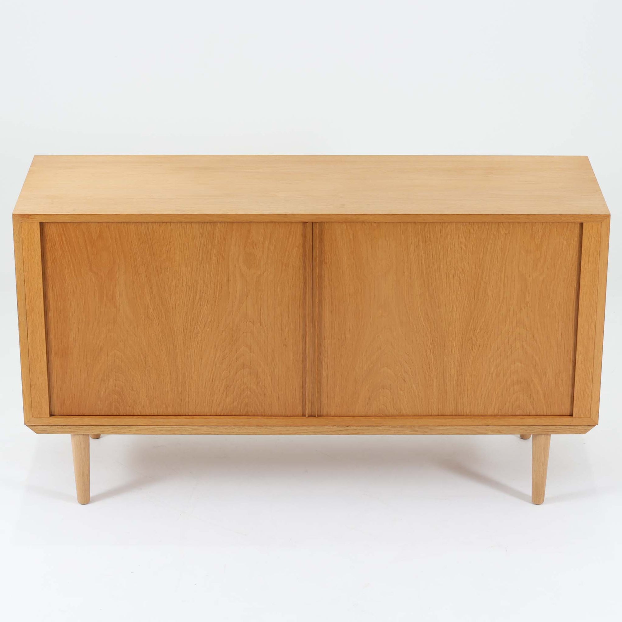 Poul Hundevadがデザインし、Hundevad &amp; Coより製作された蛇腹仕様のサイドボードです。オーク材の穏やかで上品な木目を活かした外観に、横方向へ滑らかに開閉する蛇腹扉を組み合わせた、機能美あふれる一台。扉の開閉時に前後のスペースを取らない構造は、動線を妨げず実用性にも優れています。内部は棚板構成で収納力が高く、食器や書類、リビング周りの小物まで幅広く対応。細身の脚部が全体を軽やかに引き締め、経年によって深みを増した素材感が北欧ビンテージならではの静かな存在感を空間にもたらします。