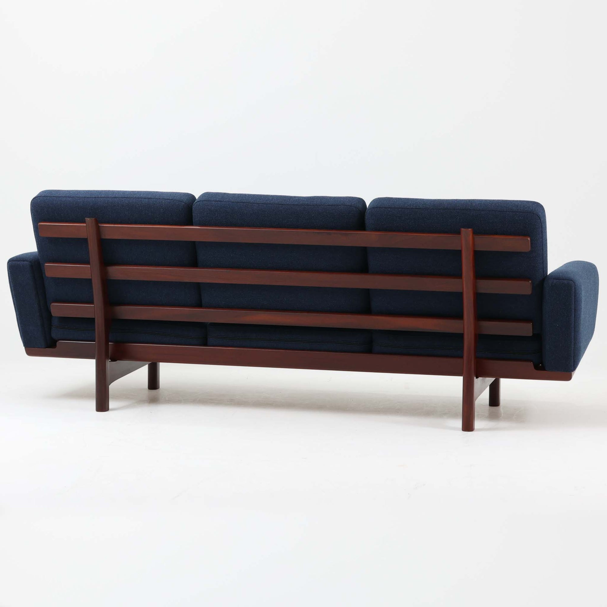 こちらは、ウェグナーデザインの人気モデル、GE236の3人掛けソファです。後ろ姿が非常に美しいため、あえて壁に寄せず背面を見せる配置もおすすめです。クッション内部にはスプリングが内蔵されており、座り心地も良好です。さらに、こちらは現地でも希少なアフロモシア材（通称アフリカンチーク）が使用された特別なモデルです。GE236のチーク材仕様として一般的に認識されているのは、このアフロモシア材が使用されたモデルを指します。GETAMA社の刻印も確認できます。