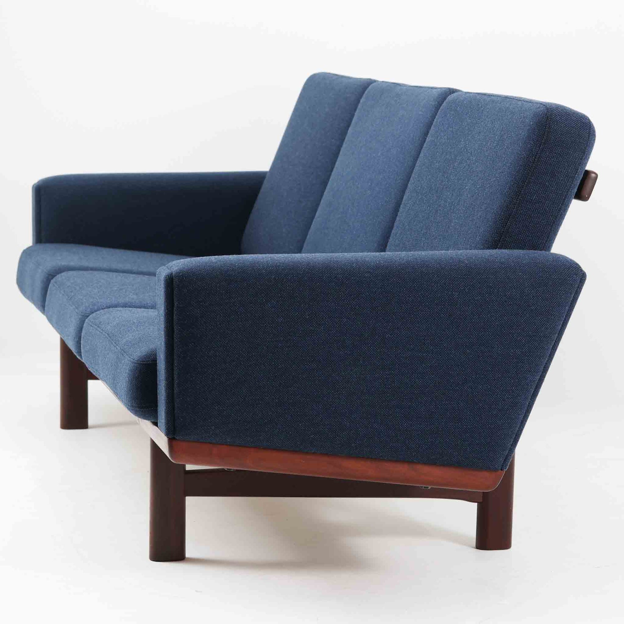 こちらは、ウェグナーデザインの人気モデル、GE236の3人掛けソファです。後ろ姿が非常に美しいため、あえて壁に寄せず背面を見せる配置もおすすめです。クッション内部にはスプリングが内蔵されており、座り心地も良好です。さらに、こちらは現地でも希少なアフロモシア材（通称アフリカンチーク）が使用された特別なモデルです。GE236のチーク材仕様として一般的に認識されているのは、このアフロモシア材が使用されたモデルを指します。GETAMA社の刻印も確認できます。