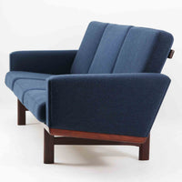 こちらは、ウェグナーデザインの人気モデル、GE236の3人掛けソファです。後ろ姿が非常に美しいため、あえて壁に寄せず背面を見せる配置もおすすめです。クッション内部にはスプリングが内蔵されており、座り心地も良好です。さらに、こちらは現地でも希少なアフロモシア材（通称アフリカンチーク）が使用された特別なモデルです。GE236のチーク材仕様として一般的に認識されているのは、このアフロモシア材が使用されたモデルを指します。GETAMA社の刻印も確認できます。