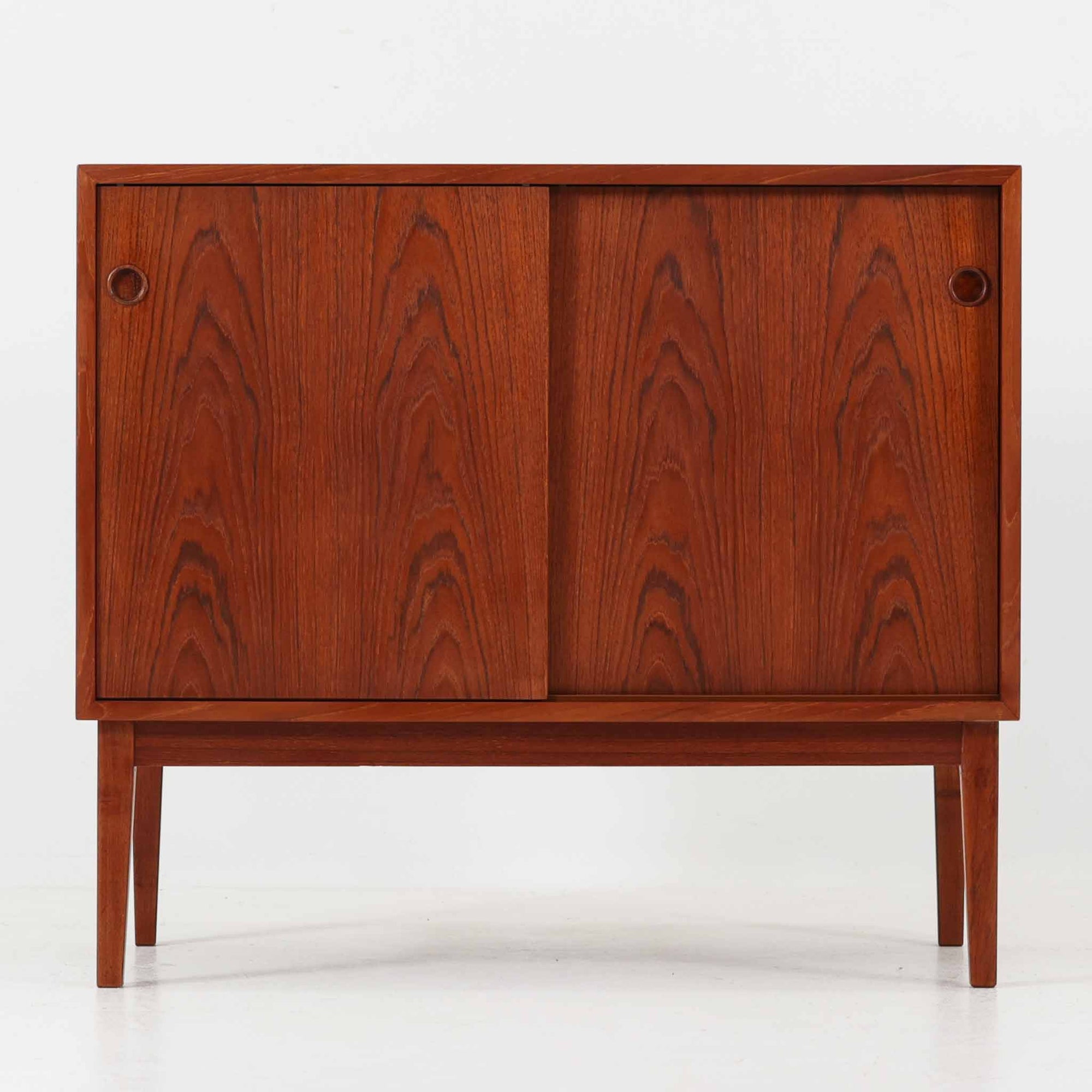 デンマーク製ローボードです。チーク材ならではの赤みを帯びた美しい木目が前面にあらわれ、引き戸には掘り込み式の丸い取っ手を配したミニマルな意匠が北欧らしい静かな存在感を放ちます。幅80cmの小ぶりなサイズ感で、リビングのテレビ台としてはもちろん、書斎やベッドルームでの収納家具としても取り入れやすく、内部には棚板を備え実用性も十分です。経年によって深みを増した木肌の表情と丁寧なつくりが、ビンテージならではの温もりを空間に添えてくれる一台です。