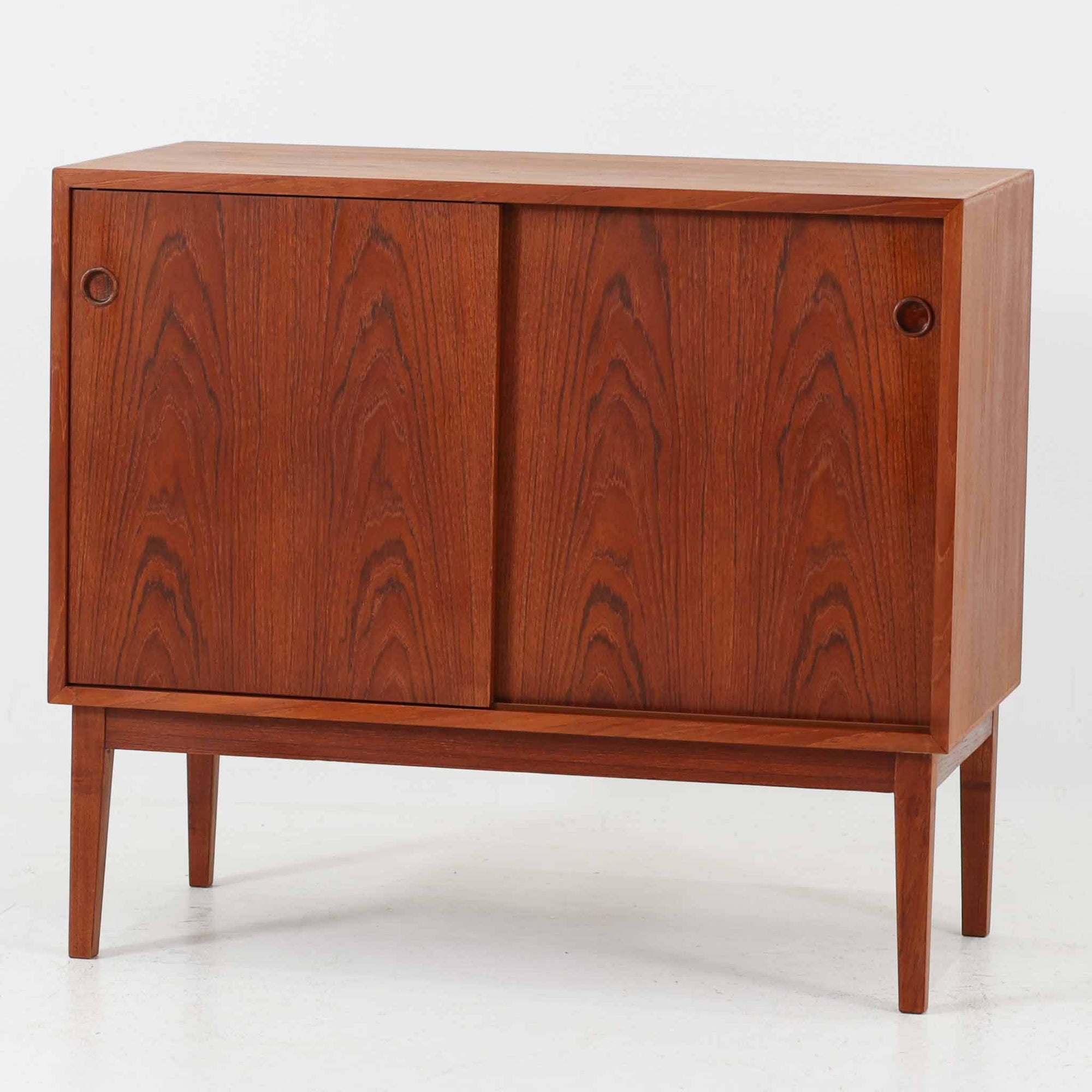 デンマーク製ローボードです。チーク材ならではの赤みを帯びた美しい木目が前面にあらわれ、引き戸には掘り込み式の丸い取っ手を配したミニマルな意匠が北欧らしい静かな存在感を放ちます。幅80cmの小ぶりなサイズ感で、リビングのテレビ台としてはもちろん、書斎やベッドルームでの収納家具としても取り入れやすく、内部には棚板を備え実用性も十分です。経年によって深みを増した木肌の表情と丁寧なつくりが、ビンテージならではの温もりを空間に添えてくれる一台です。