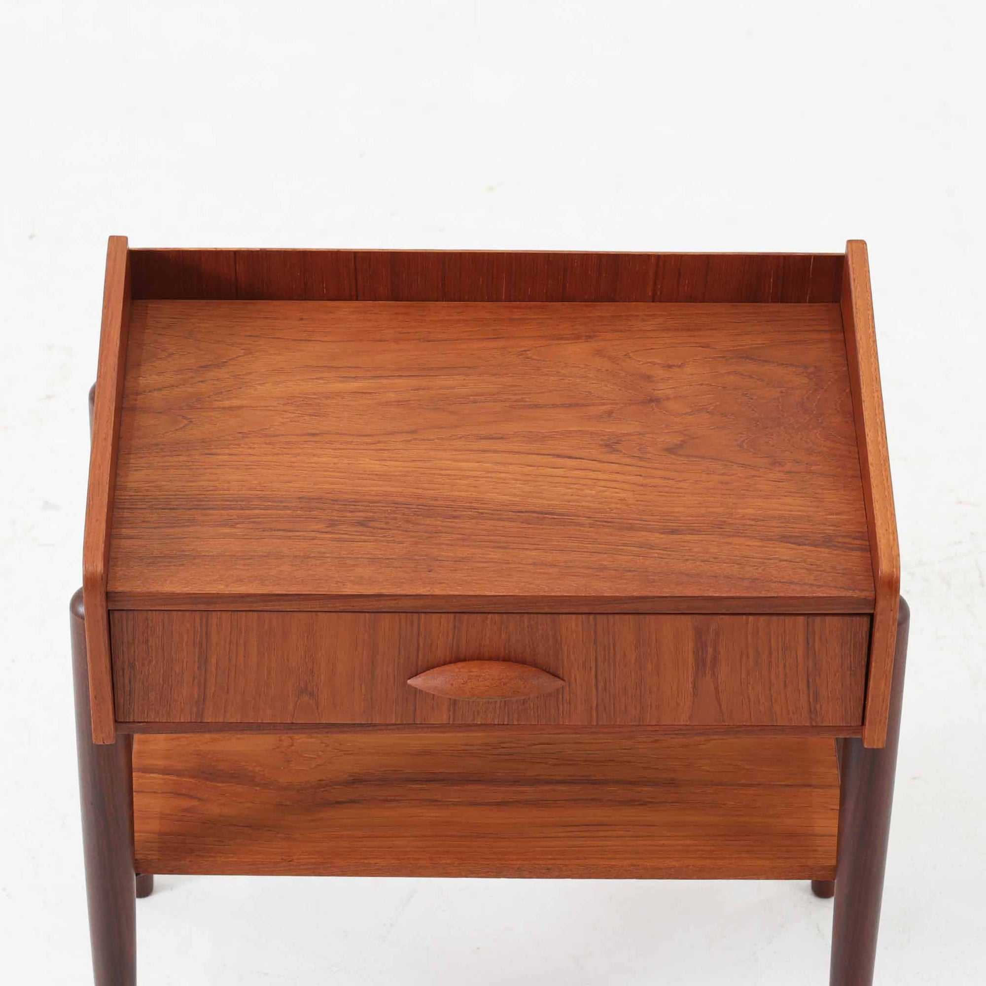 デンマーク製のサイドチェストです。チーク材ならではの温かみある木目と落ち着いた色合いが美しく、側板を立ち上げた天板形状がさりげないデザインアクセントになっています。引き出しには手掛けとして一体成形された取っ手があしらわれ、滑らかな開閉とすっきりとした正面意匠を両立しています。幅52cmの扱いやすいサイズ感で、ベッドサイドやソファ脇、玄関先などにも取り入れやすく、下段の棚板は本や小物の収納にも便利です。経年変化によって深みを増したチークの表情が、ビンテージならではの落ち着いた存在感を空間にもたらします。