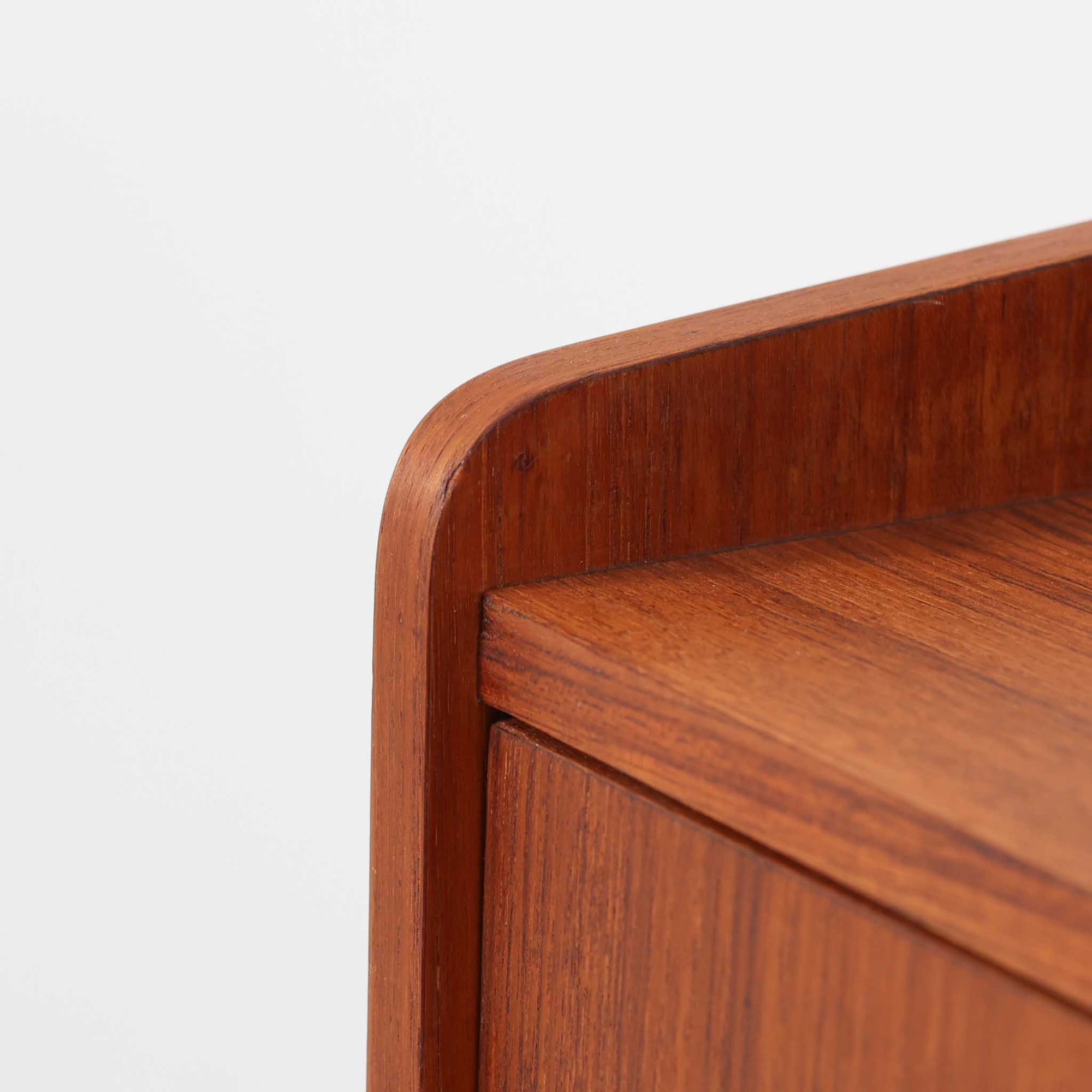 デンマーク製のサイドチェストです。チーク材ならではの温かみある木目と落ち着いた色合いが美しく、側板を立ち上げた天板形状がさりげないデザインアクセントになっています。引き出しには手掛けとして一体成形された取っ手があしらわれ、滑らかな開閉とすっきりとした正面意匠を両立しています。幅52cmの扱いやすいサイズ感で、ベッドサイドやソファ脇、玄関先などにも取り入れやすく、下段の棚板は本や小物の収納にも便利です。経年変化によって深みを増したチークの表情が、ビンテージならではの落ち着いた存在感を空間にもたらします。