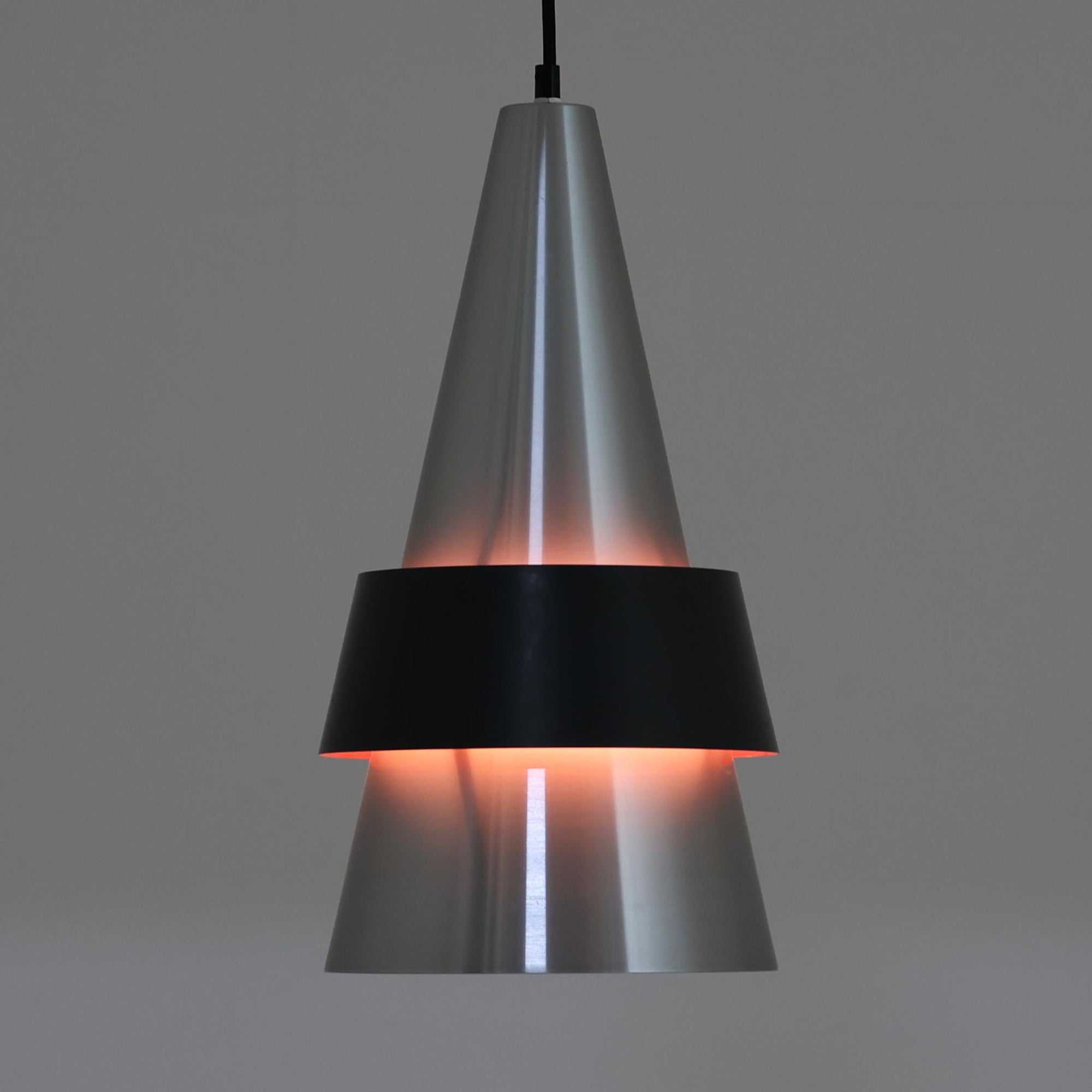 Jo HammerborgがFog & Morupのために手掛けた「Corona」は、直径22cmとコンパクトながら全高45cmと縦にしっかりとした存在感を持つアルミ×スチール製ペンダントライトで、上下に伸びる円錐形フォルムと中央のブラックバンドが彫刻的な印象を与える北欧ビンテージ品です。光源を囲む二重構造によって眩しさを抑えつつ下方向へ明かりを集中的に落とす設計となっており、ダイニングやカウンター上での1灯使用はもちろん、複数灯を並べても空間にリズムを生み出します。経年で深まった素材の風合いがハーマボーらしいモダンな佇まいを際立たせ、空間のアクセントとしても十分な存在感を発揮します。