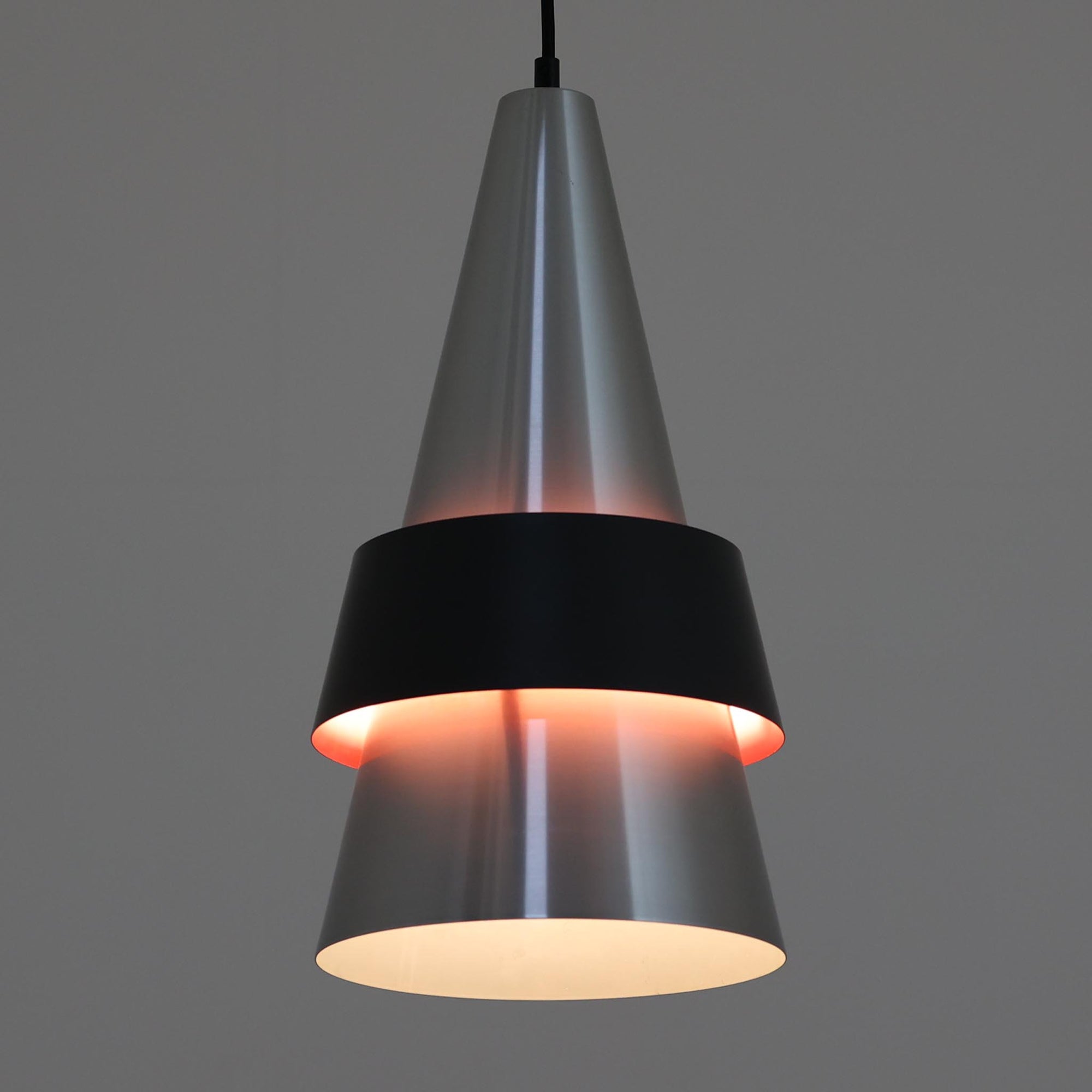 Jo HammerborgがFog &amp; Morupのために手掛けた「Corona」は、直径22cmとコンパクトながら全高45cmと縦にしっかりとした存在感を持つアルミ×スチール製ペンダントライトで、上下に伸びる円錐形フォルムと中央のブラックバンドが彫刻的な印象を与える北欧ビンテージ品です。光源を囲む二重構造によって眩しさを抑えつつ下方向へ明かりを集中的に落とす設計となっており、ダイニングやカウンター上での1灯使用はもちろん、複数灯を並べても空間にリズムを生み出します。経年で深まった素材の風合いがハーマボーらしいモダンな佇まいを際立たせ、空間のアクセントとしても十分な存在感を発揮します。