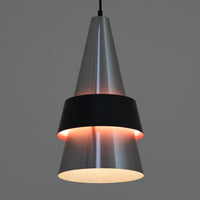 Jo HammerborgがFog & Morupのために手掛けた「Corona」は、直径22cmとコンパクトながら全高45cmと縦にしっかりとした存在感を持つアルミ×スチール製ペンダントライトで、上下に伸びる円錐形フォルムと中央のブラックバンドが彫刻的な印象を与える北欧ビンテージ品です。光源を囲む二重構造によって眩しさを抑えつつ下方向へ明かりを集中的に落とす設計となっており、ダイニングやカウンター上での1灯使用はもちろん、複数灯を並べても空間にリズムを生み出します。経年で深まった素材の風合いがハーマボーらしいモダンな佇まいを際立たせ、空間のアクセントとしても十分な存在感を発揮します。