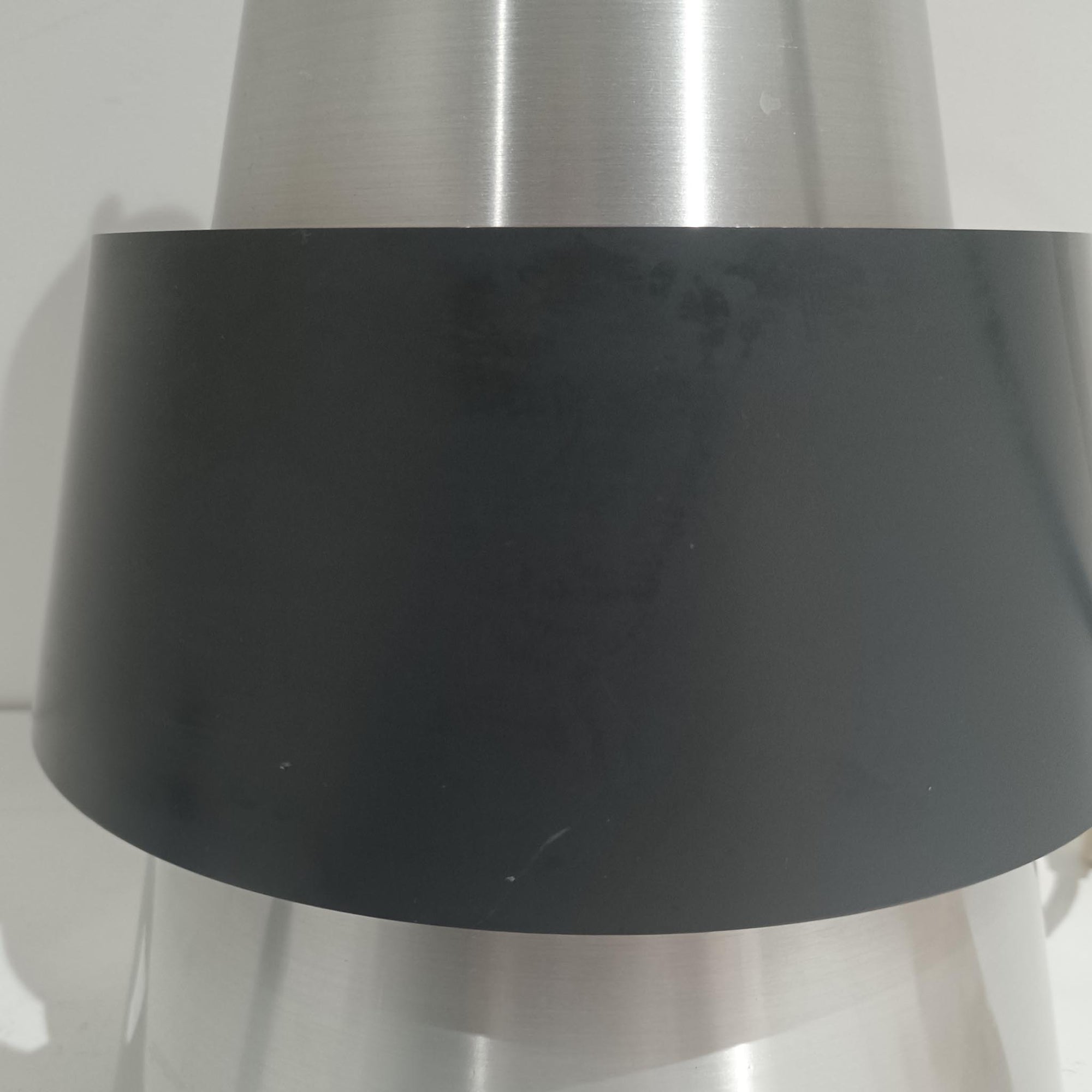 Jo HammerborgがFog &amp; Morupのために手掛けた「Corona」は、直径22cmとコンパクトながら全高45cmと縦にしっかりとした存在感を持つアルミ×スチール製ペンダントライトで、上下に伸びる円錐形フォルムと中央のブラックバンドが彫刻的な印象を与える北欧ビンテージ品です。光源を囲む二重構造によって眩しさを抑えつつ下方向へ明かりを集中的に落とす設計となっており、ダイニングやカウンター上での1灯使用はもちろん、複数灯を並べても空間にリズムを生み出します。経年で深まった素材の風合いがハーマボーらしいモダンな佇まいを際立たせ、空間のアクセントとしても十分な存在感を発揮します。