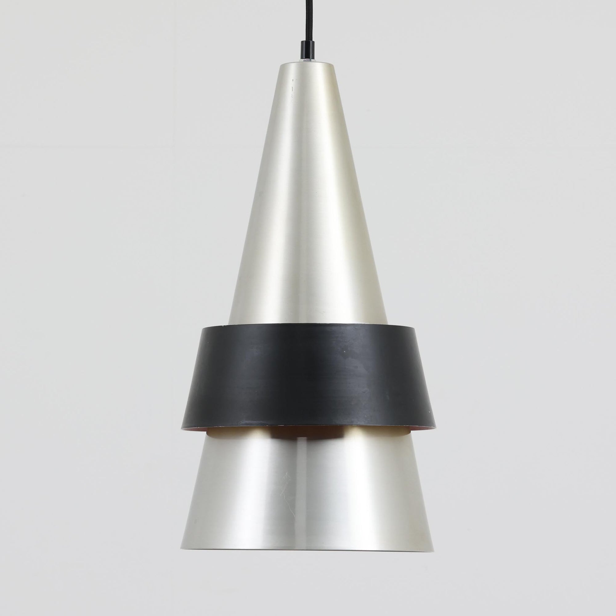 Jo HammerborgがFog & Morupのために手掛けた「Corona」は、直径22cmとコンパクトながら全高45cmと縦にしっかりとした存在感を持つアルミ×スチール製ペンダントライトで、上下に伸びる円錐形フォルムと中央のブラックバンドが彫刻的な印象を与える北欧ビンテージ品です。光源を囲む二重構造によって眩しさを抑えつつ下方向へ明かりを集中的に落とす設計となっており、ダイニングやカウンター上での1灯使用はもちろん、複数灯を並べても空間にリズムを生み出します。経年で深まった素材の風合いがハーマボーらしいモダンな佇まいを際立たせ、空間のアクセントとしても十分な存在感を発揮します。