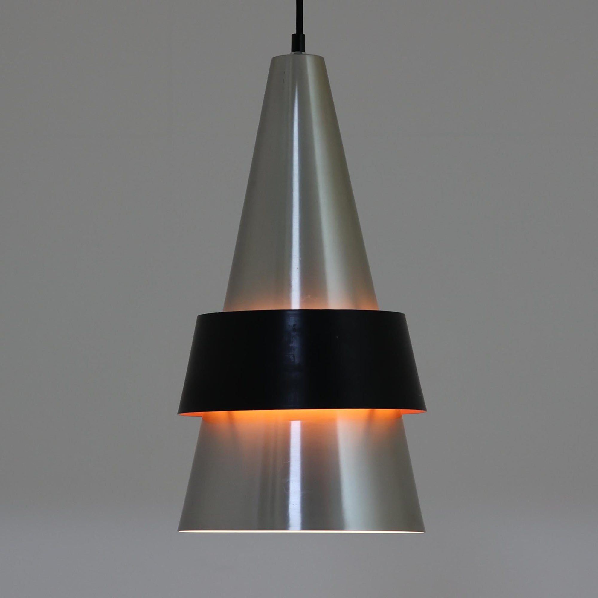 Jo HammerborgがFog & Morupのために手掛けた「Corona」は、直径22cmとコンパクトながら全高45cmと縦にしっかりとした存在感を持つアルミ×スチール製ペンダントライトで、上下に伸びる円錐形フォルムと中央のブラックバンドが彫刻的な印象を与える北欧ビンテージ品です。光源を囲む二重構造によって眩しさを抑えつつ下方向へ明かりを集中的に落とす設計となっており、ダイニングやカウンター上での1灯使用はもちろん、複数灯を並べても空間にリズムを生み出します。経年で深まった素材の風合いがハーマボーらしいモダンな佇まいを際立たせ、空間のアクセントとしても十分な存在感を発揮します。