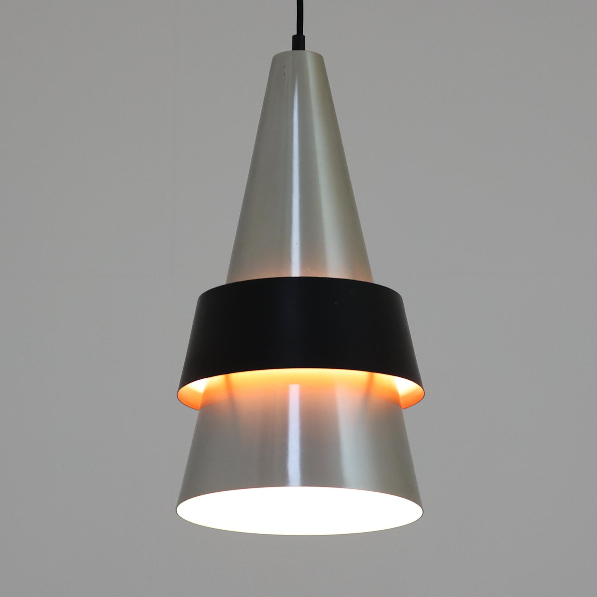 Jo HammerborgがFog &amp; Morupのために手掛けた「Corona」は、直径22cmとコンパクトながら全高45cmと縦にしっかりとした存在感を持つアルミ×スチール製ペンダントライトで、上下に伸びる円錐形フォルムと中央のブラックバンドが彫刻的な印象を与える北欧ビンテージ品です。光源を囲む二重構造によって眩しさを抑えつつ下方向へ明かりを集中的に落とす設計となっており、ダイニングやカウンター上での1灯使用はもちろん、複数灯を並べても空間にリズムを生み出します。経年で深まった素材の風合いがハーマボーらしいモダンな佇まいを際立たせ、空間のアクセントとしても十分な存在感を発揮します。