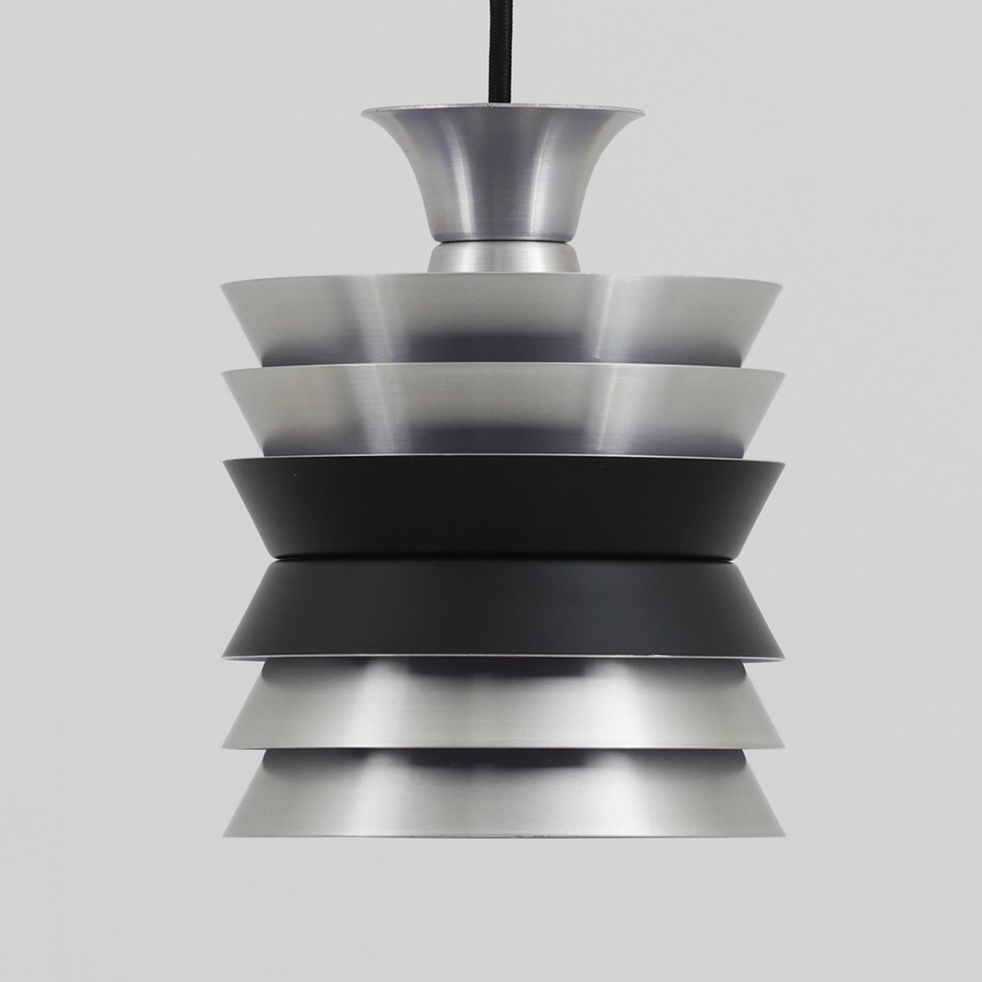 Carl ThoreがGranhaga Metallindustriで手掛けた直径15cmの小ぶりなペンダントライトで、段状に重ねたアルミシェードが光を柔らかく拡散し、点灯時には上下方向へ美しいグラデーションを生み出す北欧ビンテージ品です。繊細なヘアライン仕上げとブラックパーツのコントラストがモダンな印象を与え、キッチンカウンターや小空間のアクセント照明としてはもちろん、複数灯を並べたコーディネートにも最適です。コンパクトながら彫刻的なフォルムが存在感を放ち、経年で深まった質感がビンテージならではの魅力を際立たせています。