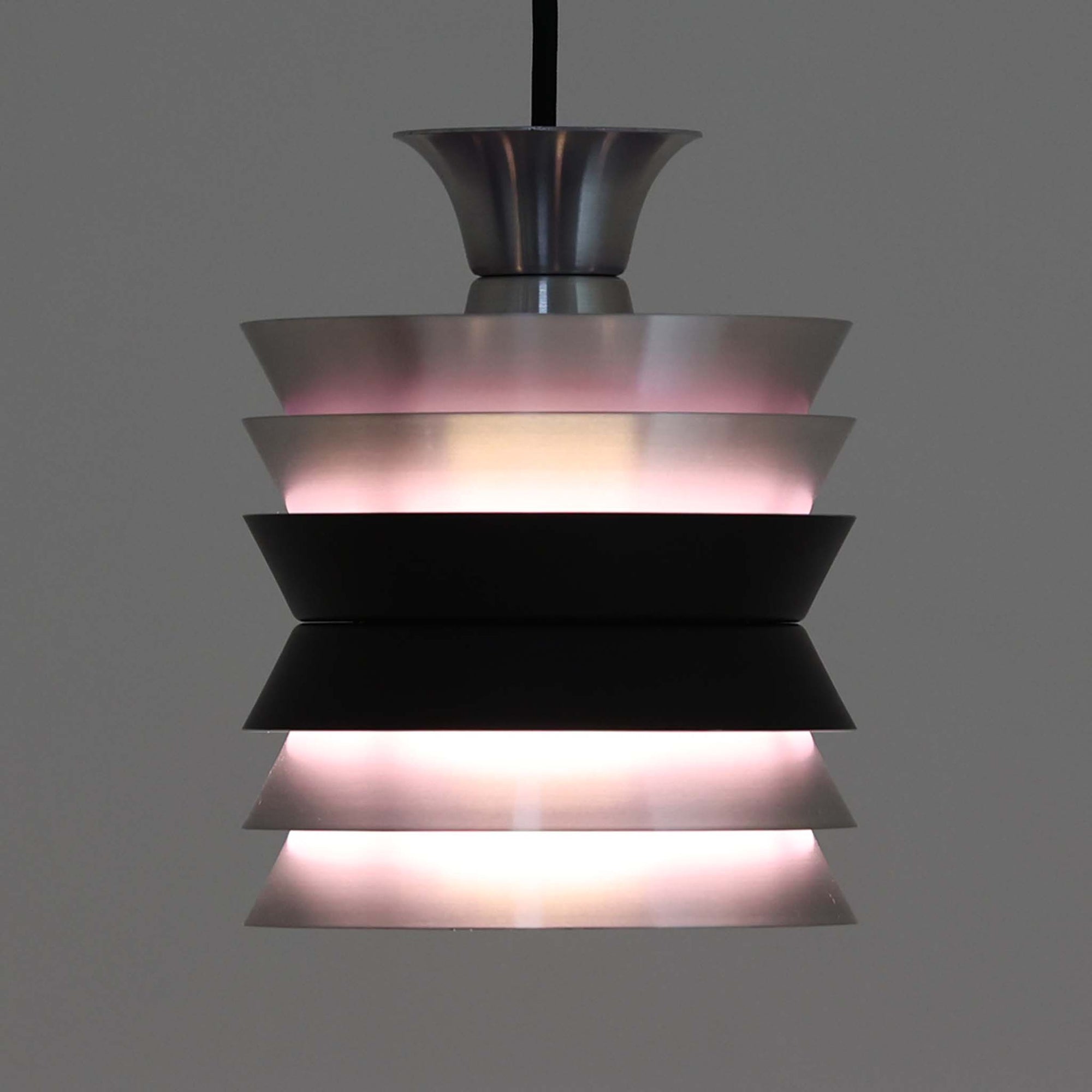 Carl ThoreがGranhaga Metallindustriで手掛けた直径15cmの小ぶりなペンダントライトで、段状に重ねたアルミシェードが光を柔らかく拡散し、点灯時には上下方向へ美しいグラデーションを生み出す北欧ビンテージ品です。繊細なヘアライン仕上げとブラックパーツのコントラストがモダンな印象を与え、キッチンカウンターや小空間のアクセント照明としてはもちろん、複数灯を並べたコーディネートにも最適です。コンパクトながら彫刻的なフォルムが存在感を放ち、経年で深まった質感がビンテージならではの魅力を際立たせています。