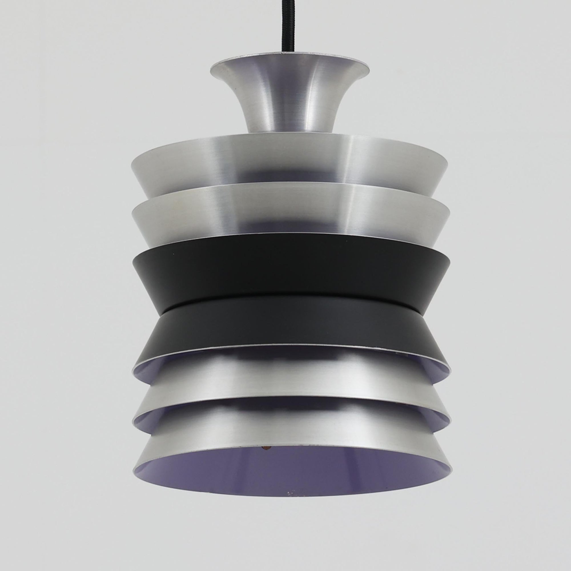 Carl ThoreがGranhaga Metallindustriで手掛けた直径15cmの小ぶりなペンダントライトで、段状に重ねたアルミシェードが光を柔らかく拡散し、点灯時には上下方向へ美しいグラデーションを生み出す北欧ビンテージ品です。繊細なヘアライン仕上げとブラックパーツのコントラストがモダンな印象を与え、キッチンカウンターや小空間のアクセント照明としてはもちろん、複数灯を並べたコーディネートにも最適です。コンパクトながら彫刻的なフォルムが存在感を放ち、経年で深まった質感がビンテージならではの魅力を際立たせています。