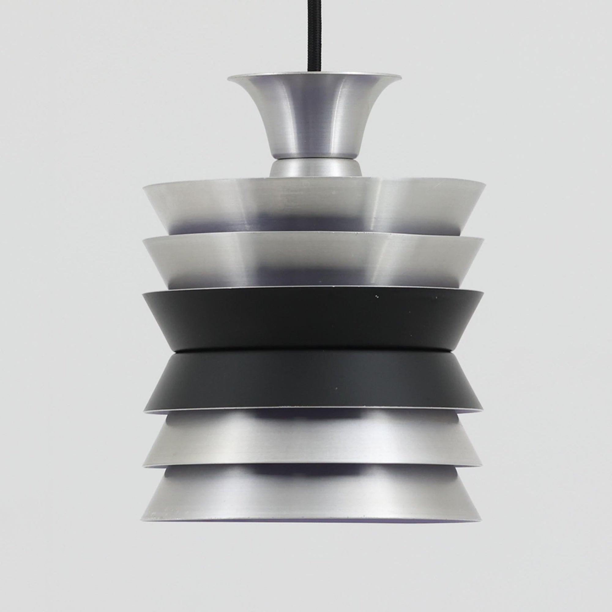 Carl ThoreがGranhaga Metallindustriで手掛けた直径15cmの小ぶりなペンダントライトで、段状に重ねたアルミシェードが光を柔らかく拡散し、点灯時には上下方向へ美しいグラデーションを生み出す北欧ビンテージ品です。繊細なヘアライン仕上げとブラックパーツのコントラストがモダンな印象を与え、キッチンカウンターや小空間のアクセント照明としてはもちろん、複数灯を並べたコーディネートにも最適です。コンパクトながら彫刻的なフォルムが存在感を放ち、経年で深まった質感がビンテージならではの魅力を際立たせています。