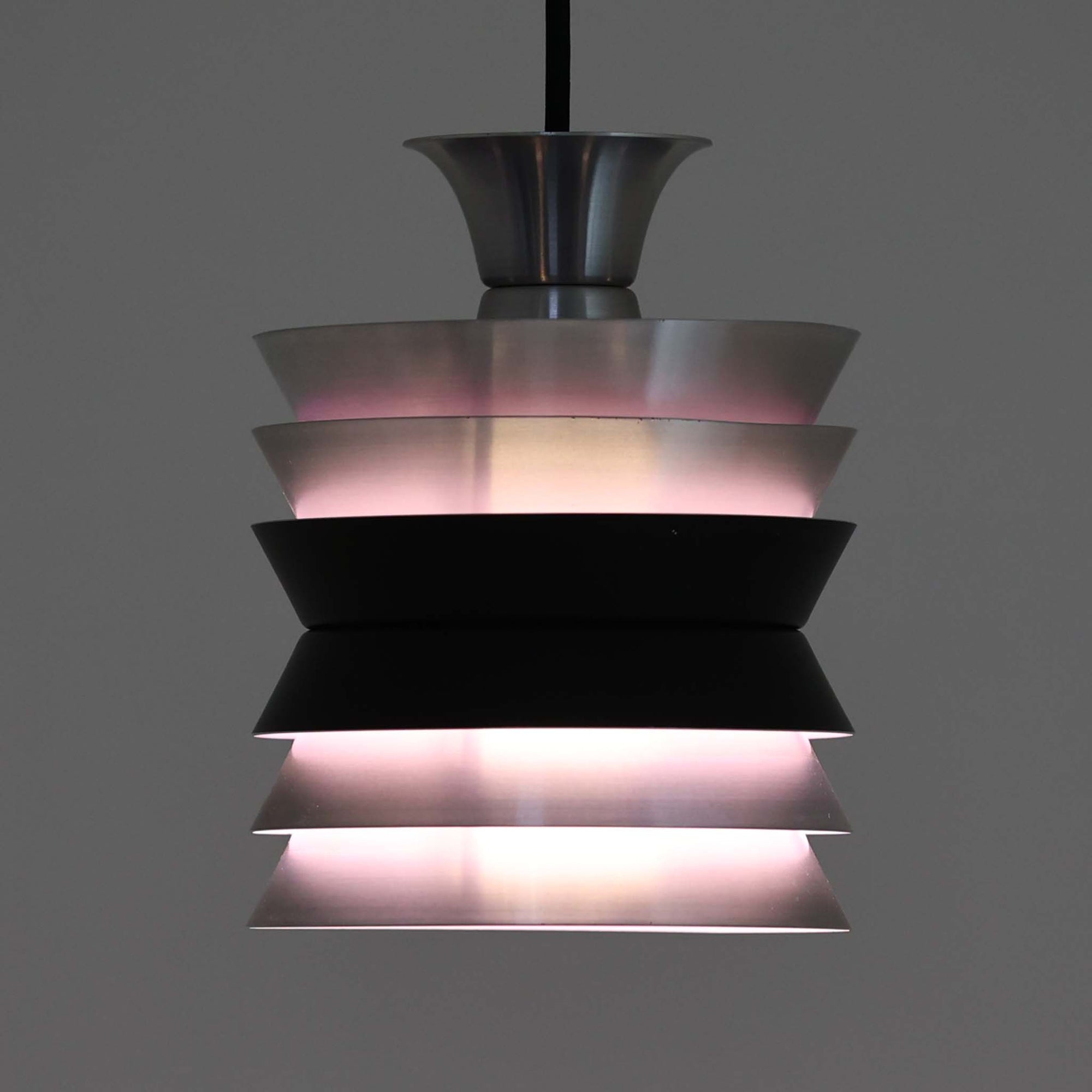 Carl ThoreがGranhaga Metallindustriで手掛けた直径15cmの小ぶりなペンダントライトで、段状に重ねたアルミシェードが光を柔らかく拡散し、点灯時には上下方向へ美しいグラデーションを生み出す北欧ビンテージ品です。繊細なヘアライン仕上げとブラックパーツのコントラストがモダンな印象を与え、キッチンカウンターや小空間のアクセント照明としてはもちろん、複数灯を並べたコーディネートにも最適です。コンパクトながら彫刻的なフォルムが存在感を放ち、経年で深まった質感がビンテージならではの魅力を際立たせています。